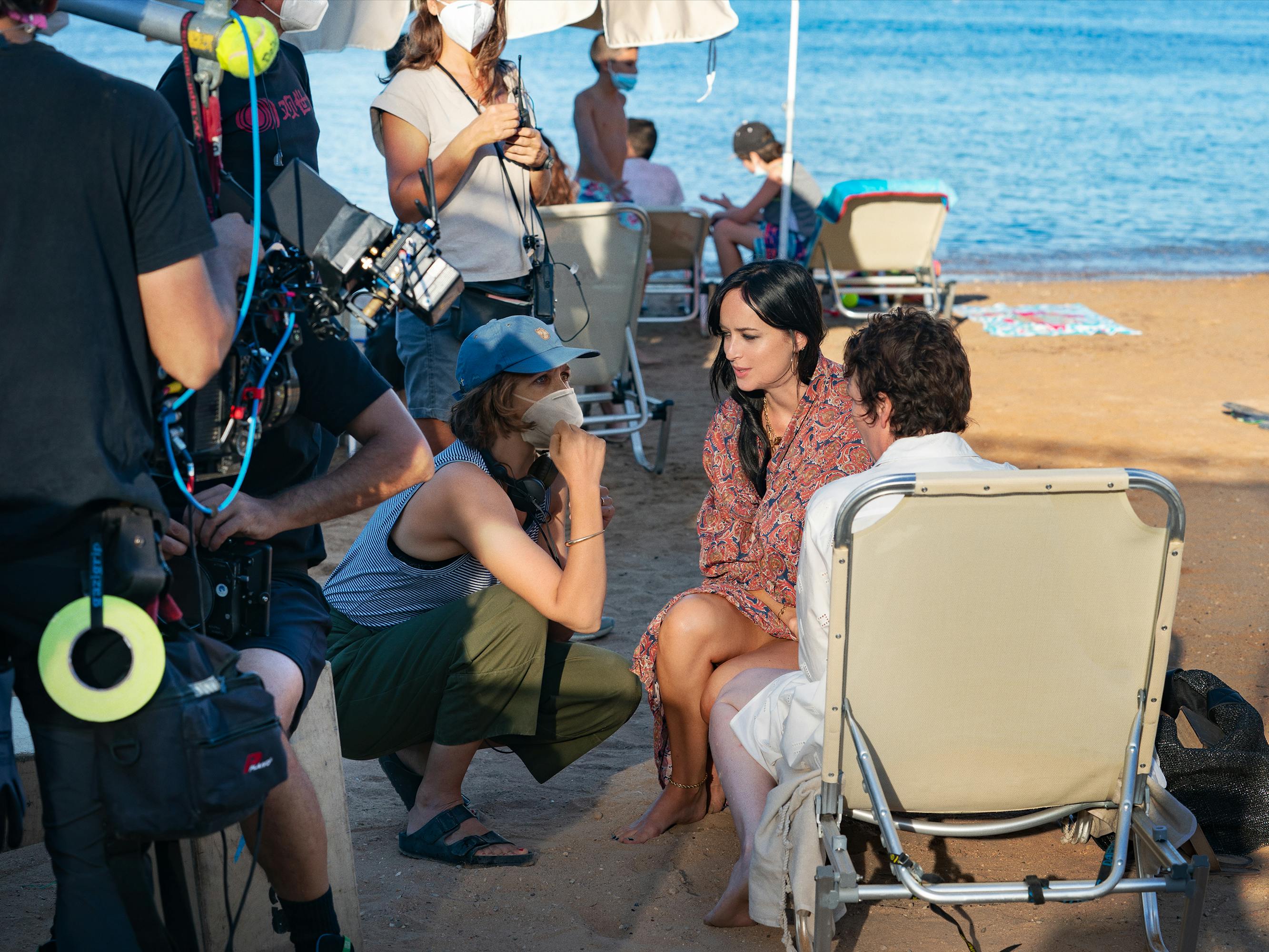 Maggie Gyllenhaal, Dakota Johnson, Olivia Colman, and The Lost Daughter crew sit on the beach. The water is a clear blue, the beach chair is creme. Colman wears a white robe, Johnson wears a red patterned shawl, and Gyllenhaal wears a blue hat and matching blue tank top.