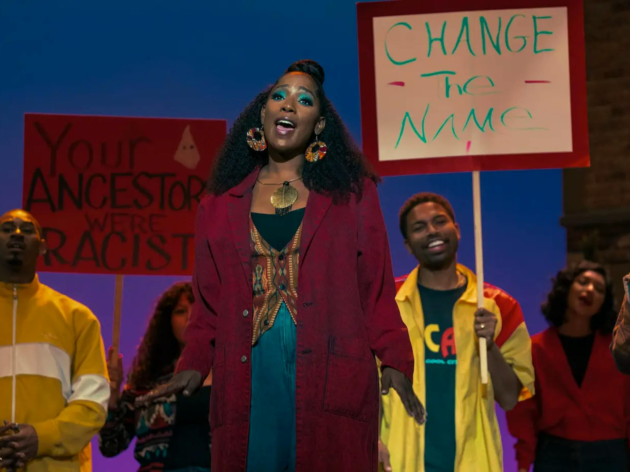 Joelle Brooks (Ashley Blaine Featherson) wears a red coat as she leads a group of people holding signs that read: ‘Your ancestors were racist’ and ‘change the name.’
