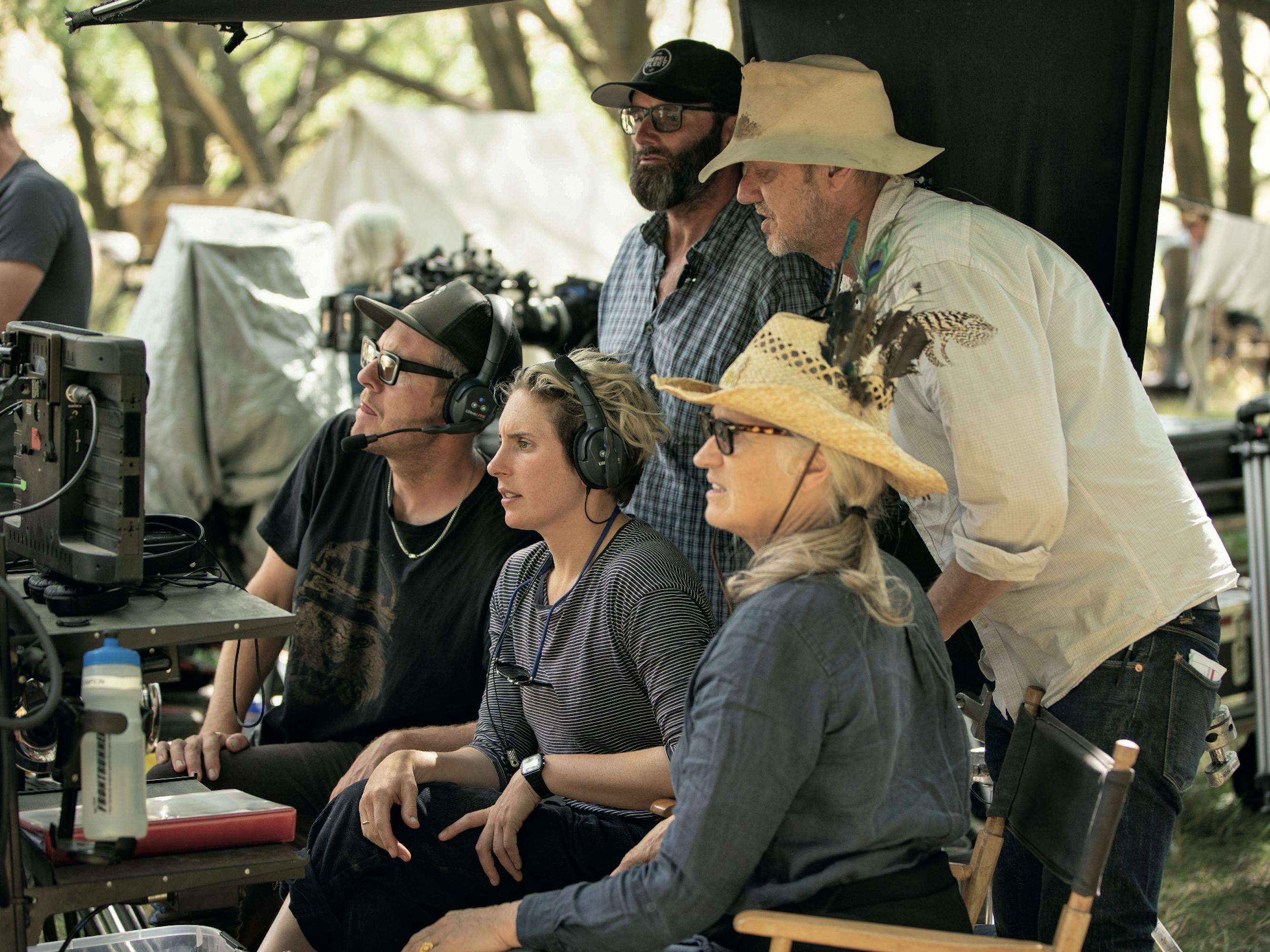 Jane Campion and The Power of the Dog crew stand together looking at something off camera. Campion wears a straw hat, grey shirt, and chunky glasses.