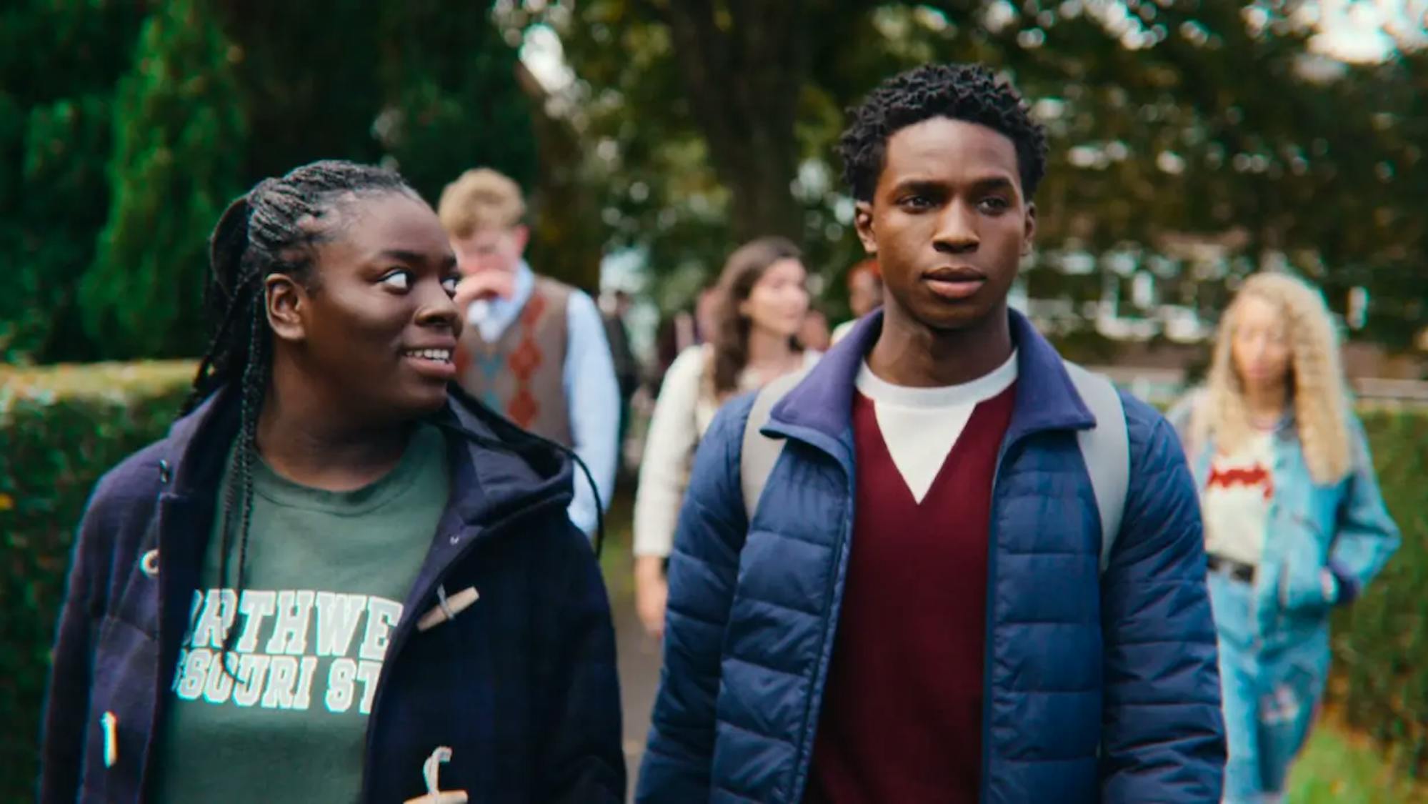 Viv Odesanya (Chinenye Edeuzu) and Jackson Marchetti (Kedar Williams-Stirling) walk together along a hedged path. She wears a green t-shirt and he wears a red sweater. They both wear navy jackets. 