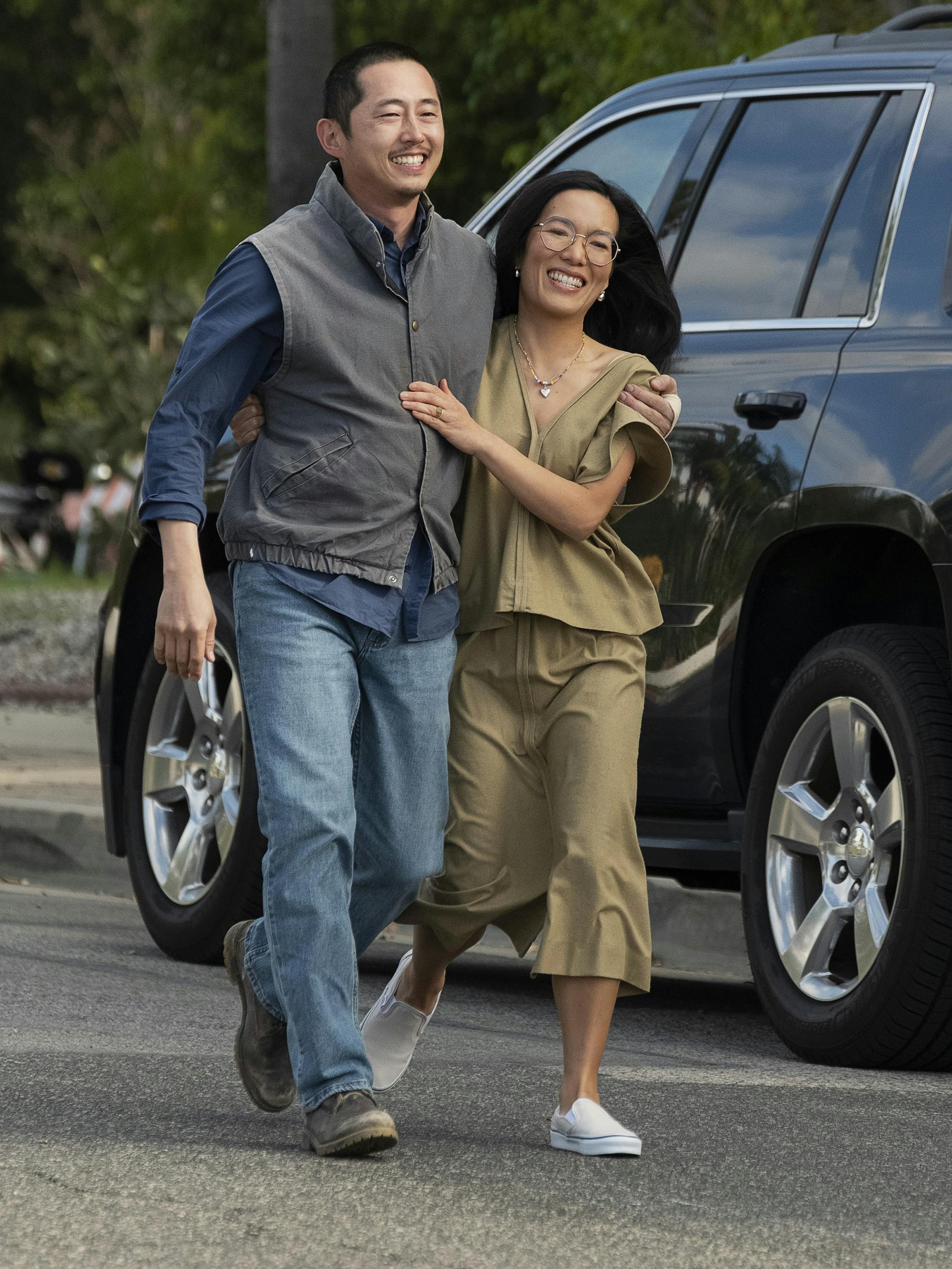 Steven Yeun and Ali Wong walk together on the set of BEEF. Yeun wears jeans and a grey vest and Wong wears a khaki matching set.