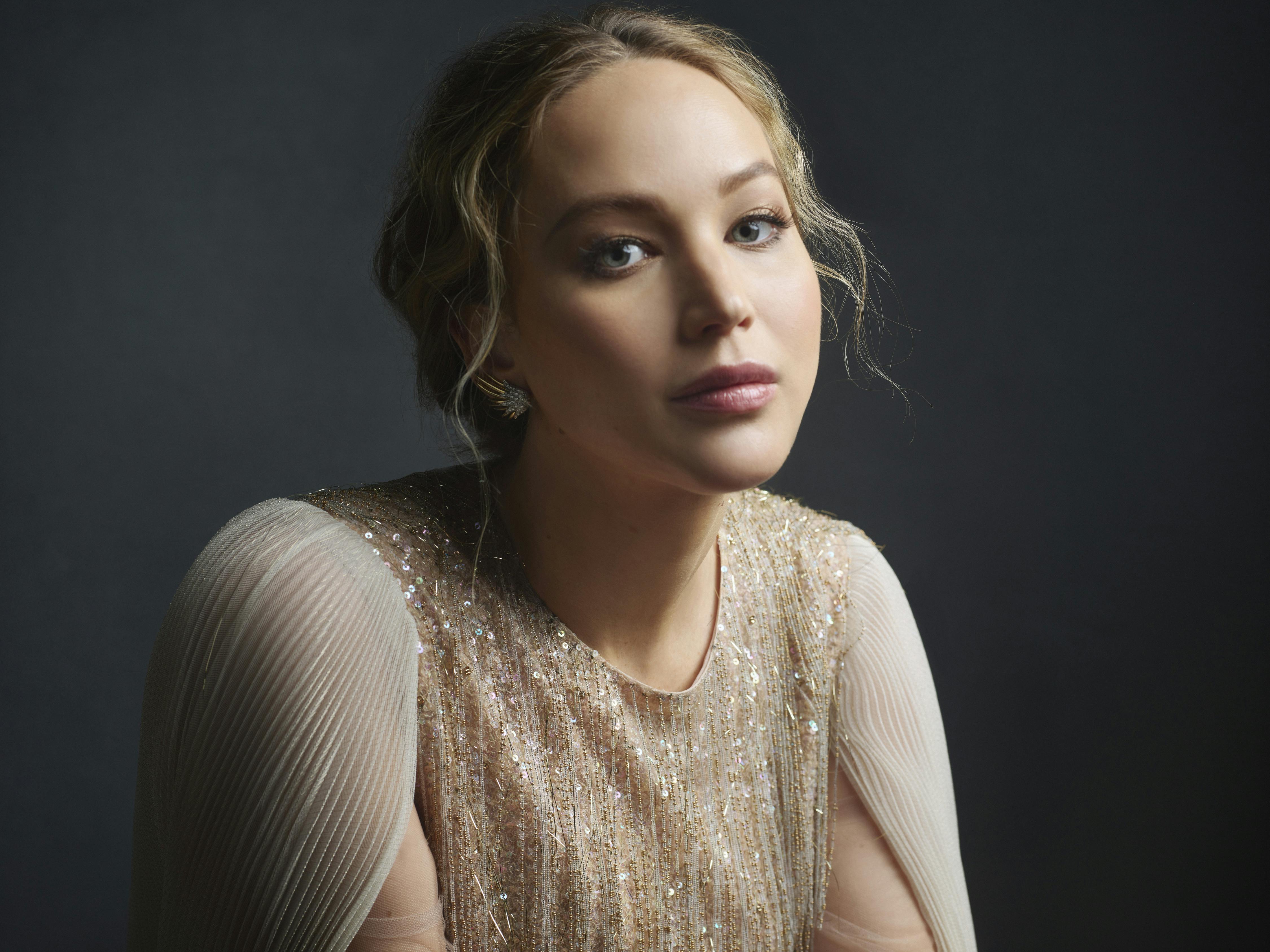 Jennifer Lawrence wears a beige sequined top. Her hair is tied back with some wispy curls framing her face.