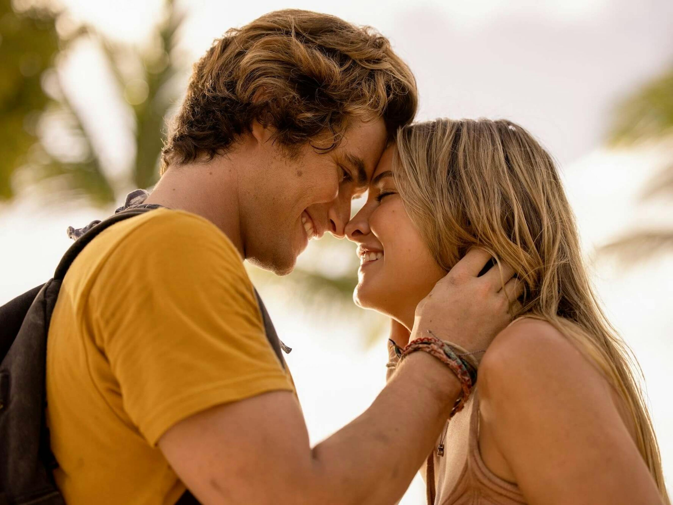 John B (Chase Stokes) and Sarah Cameron (Madelyn Cline) in Outer Banks. Chase wears a yellow t-shirt and Madelyn wears a tan tank top. They embrace with their foreheads and noses touching.