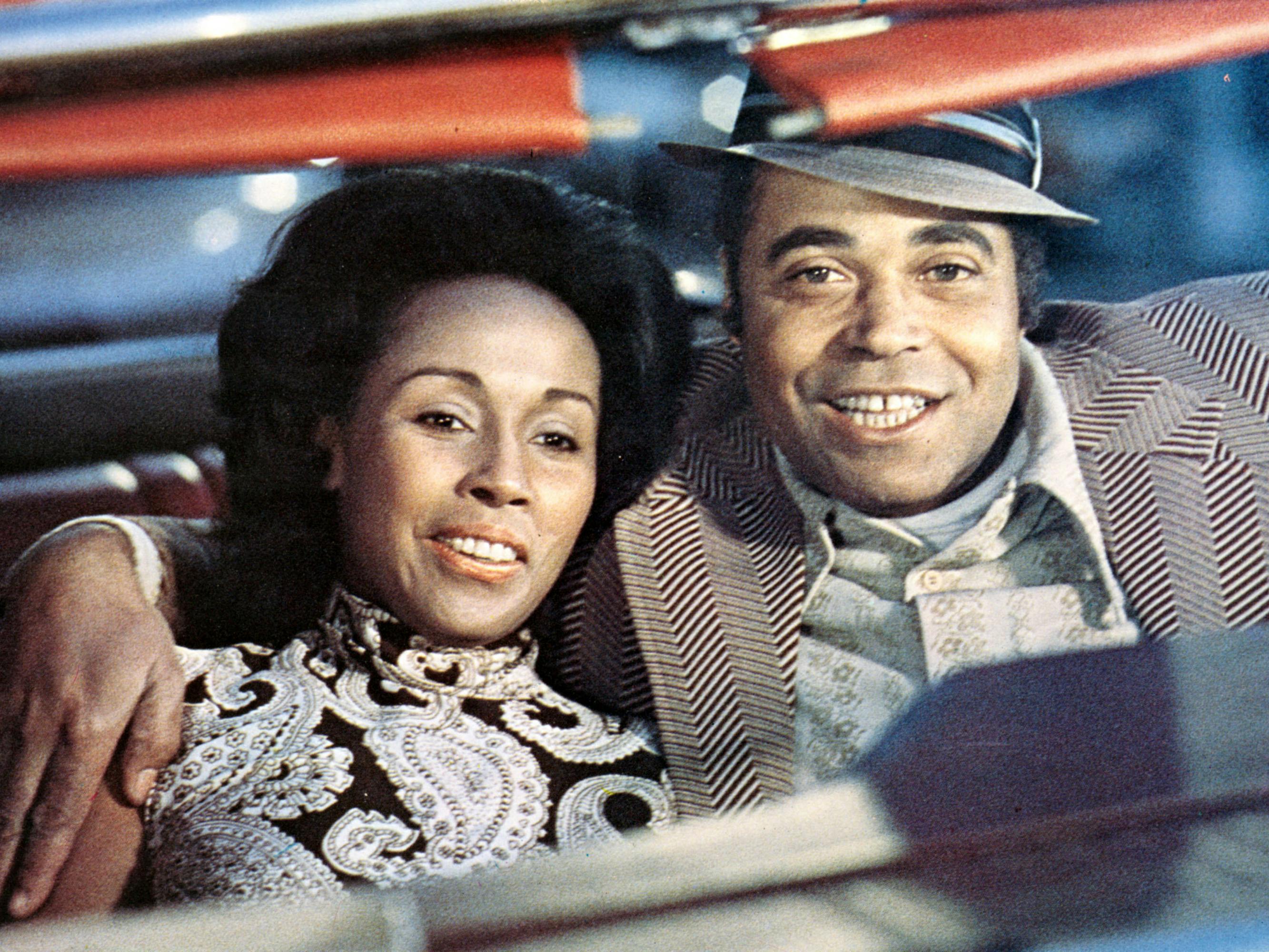 Claudine (Diahann Carroll) and Roop (James Earl Jones) sit together smiling in a car.