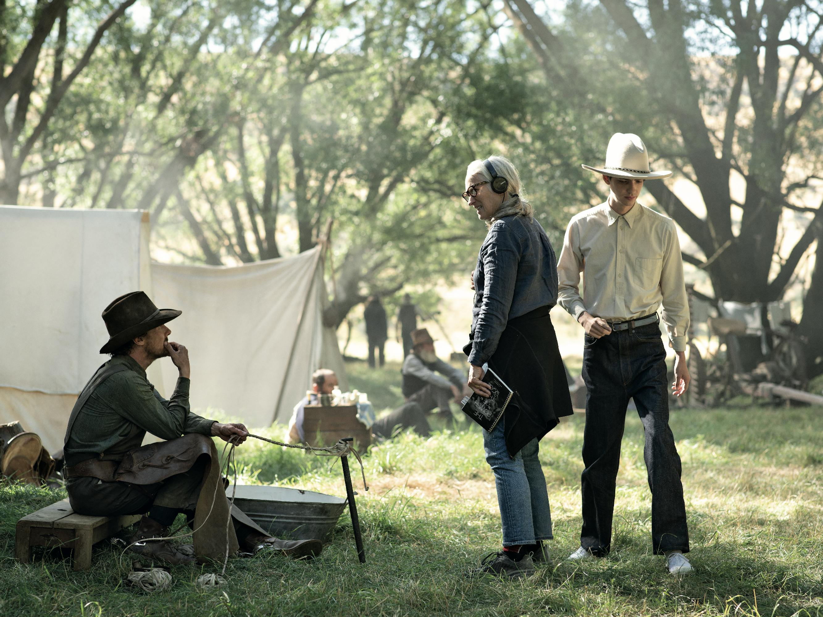 Benedict Cumberbatch, Jane Campion, and Kodi Smit-McPhee mingle outside in this green, sun-lit scene. Cumberbatch wears a wide-brimmed hat and dark clothing. Campion wears headphones, jeans, and a grey top. Smit-McPhee wears a white wide brimmed hat and dark pants.