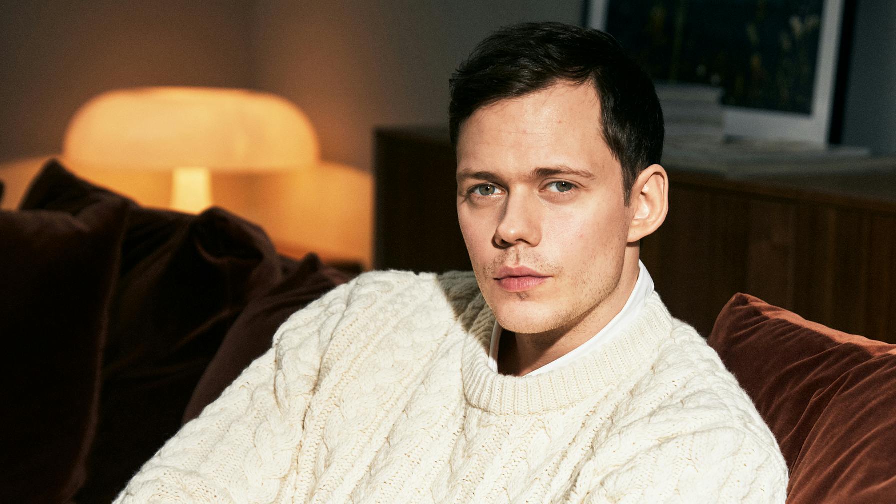 Bill Skarsgård wears a white chunky sweater and sits on a velvet brown couch.