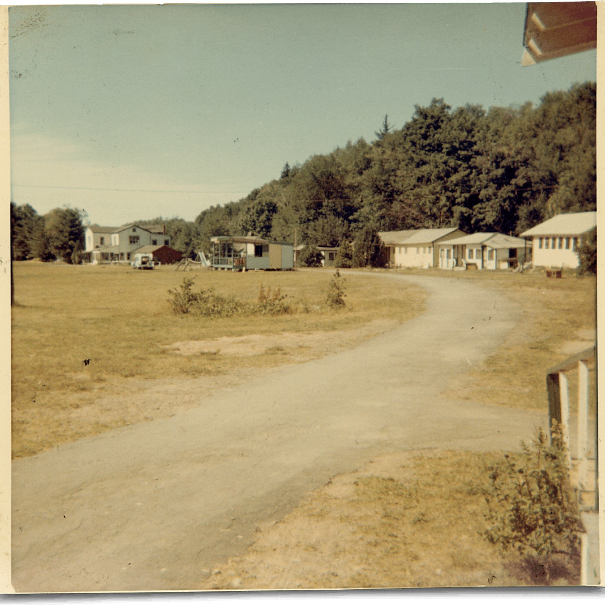 Camp Jened is pictured in a sun-faded photograph. Low, white buildings line the paved road through camp, and trees rise up behind them.