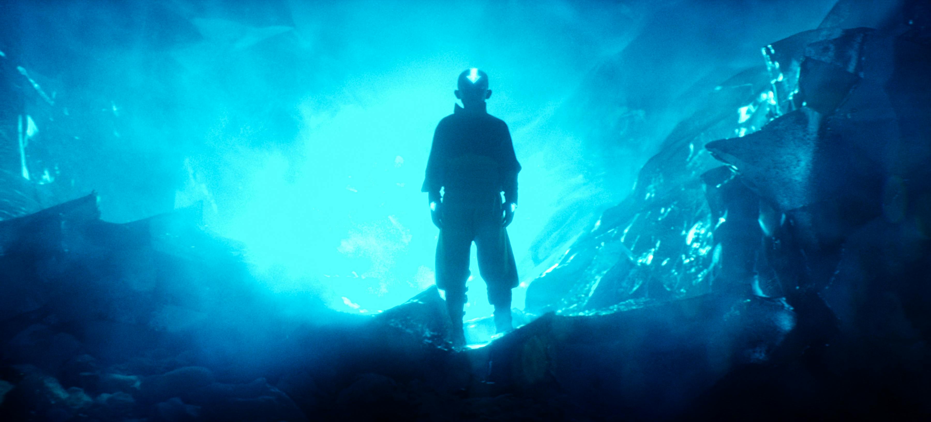 Aang (Gordon Cormier) stands atop rolling waves, backlit by neon blue light. 