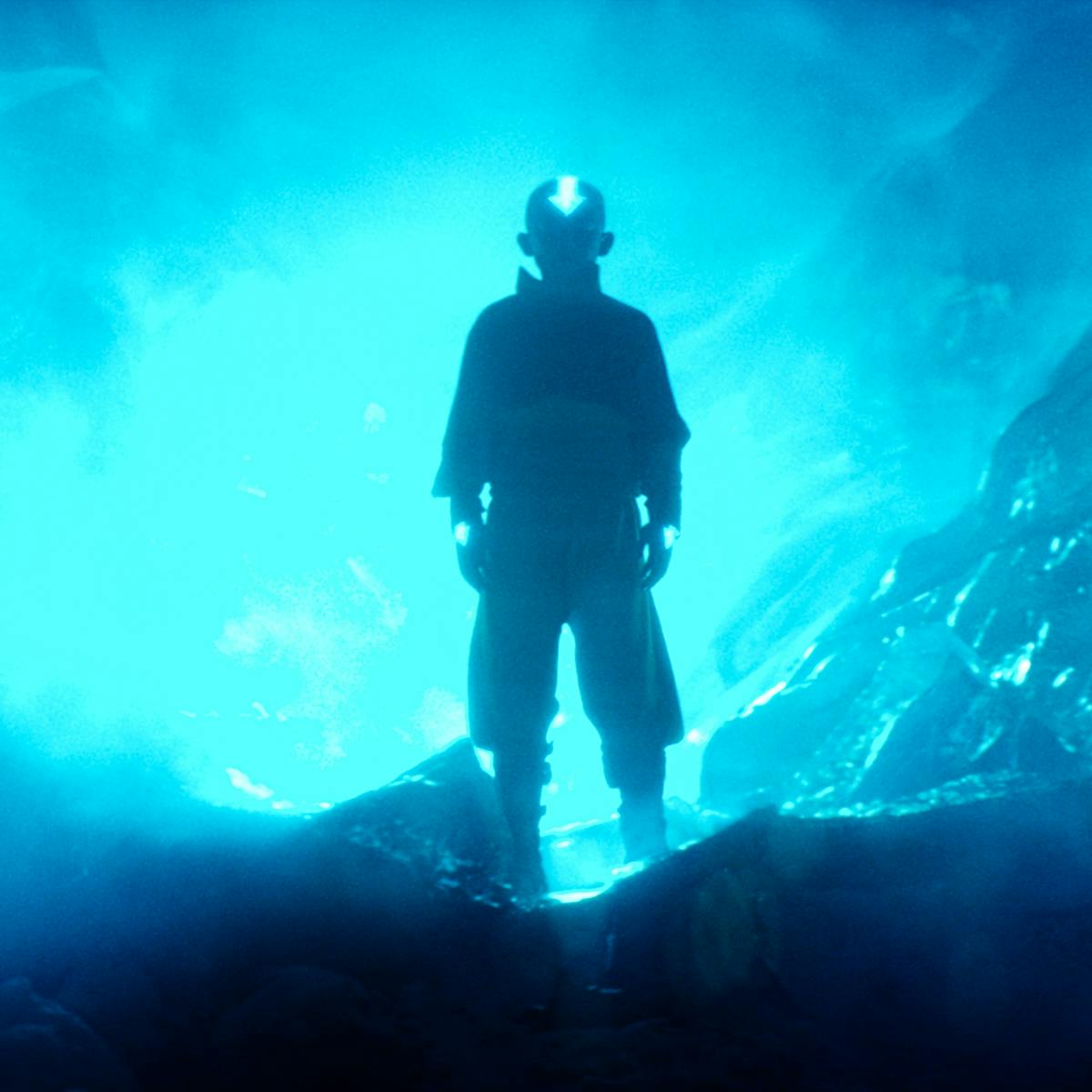 Aang (Gordon Cormier) stands atop rolling waves, backlit by neon blue light. 