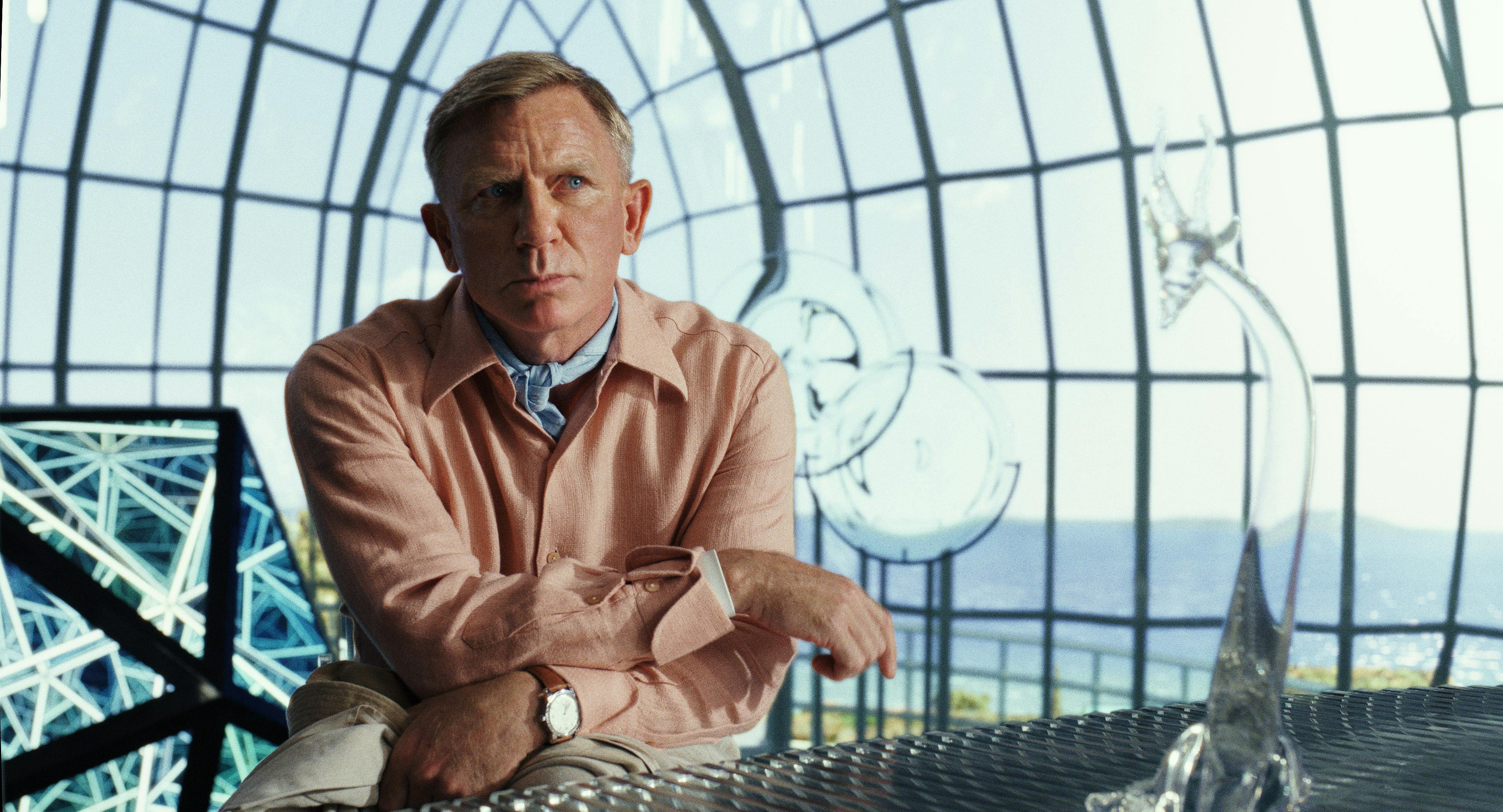 Benoit Blanc wears a light pink jacket and light blue ascot, and looks puzzled in the glass onion overlooking the sea.