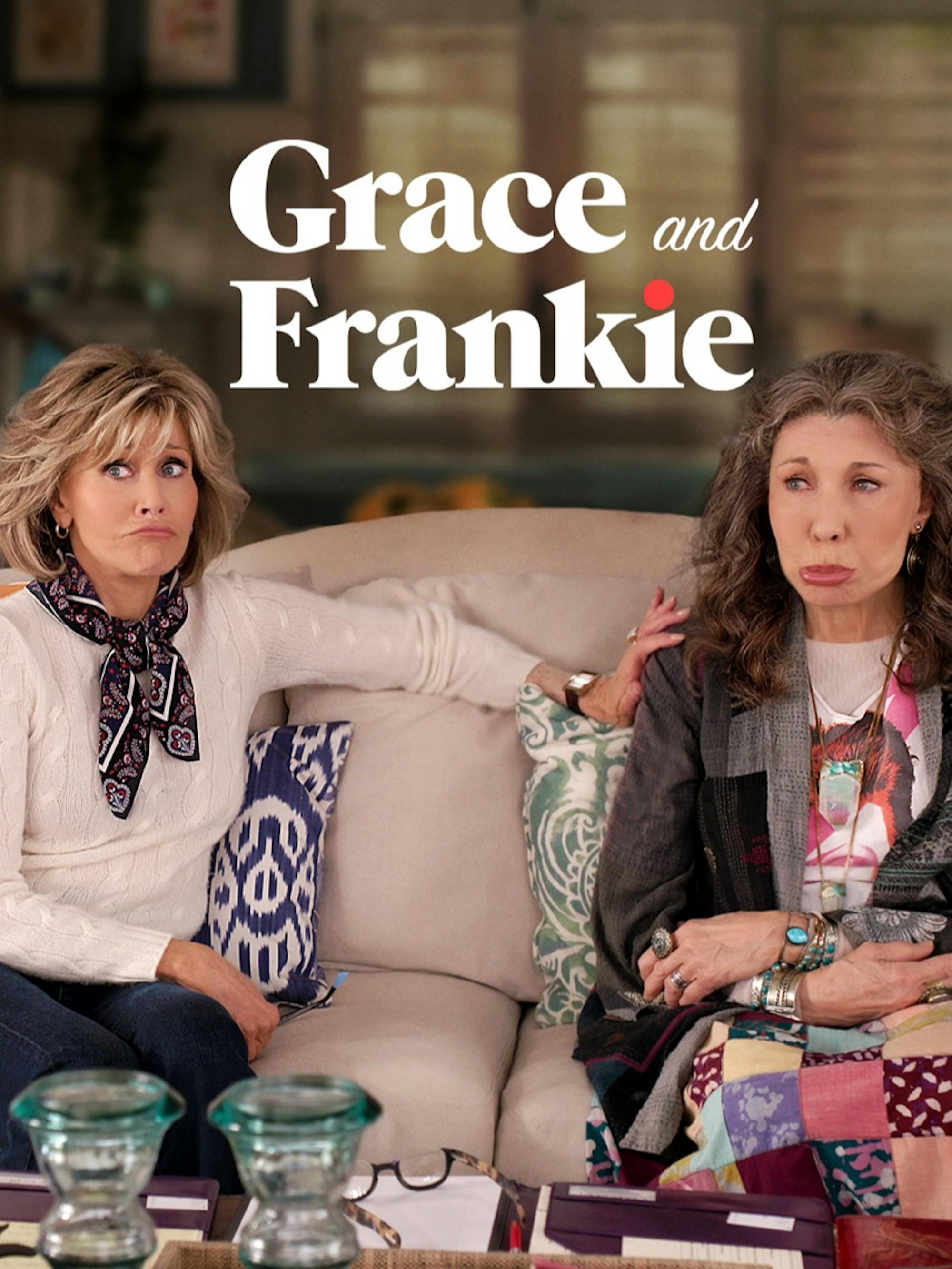 Grace and Frankie sit on a beige couch with multi colored pillows, making silly pouting faces. There are books, reading glasses, and candlestick holders in front of them, and they are dressed in their quintessential, although very different, style. 