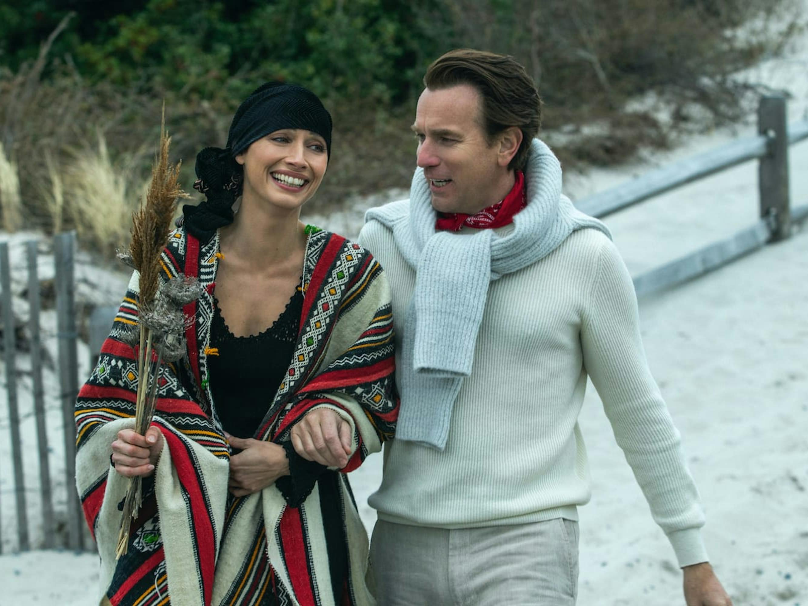 Elsa Peretti (Rebecca Dayan) and Halston (Ewan McGregor) walk on a chilly beach. Halston looks unrecognizable in his all white outfit, with a sweater tied around his shoulders. Elsa Peretti wears a scarf like cover, with a black scarf tied around her head. They laugh in this happy candid moment.