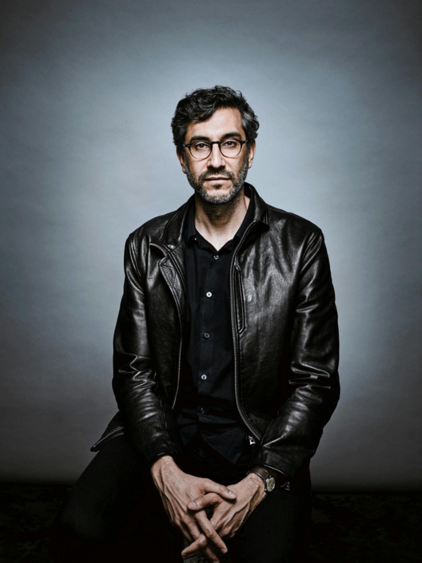 Ramin Bahrani is photographed against a gray backdrop. He is in all black, including a leather jacket, and has his hands laced on his lap. 