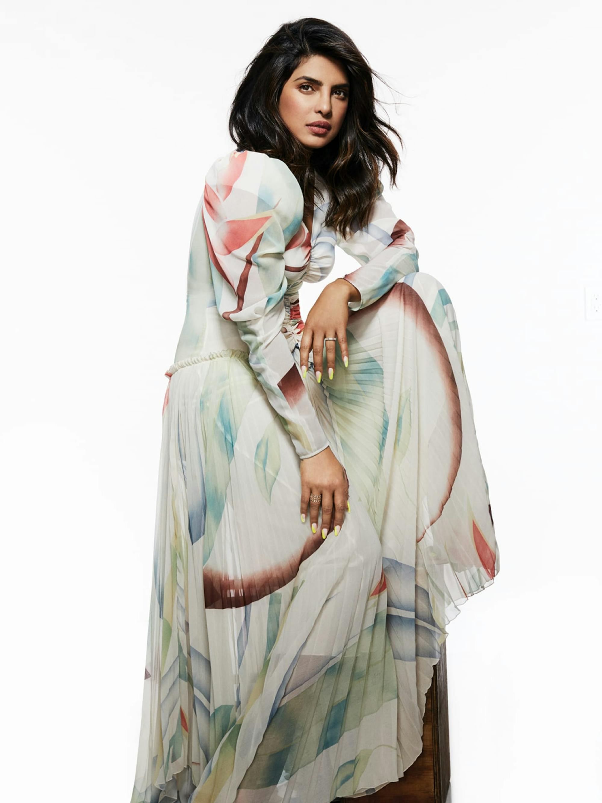 Priyanka Chopra Jonas poses perched on a seat. The skirt of her colorful dress cascades like a waterfall. Her manicure is impeccable.