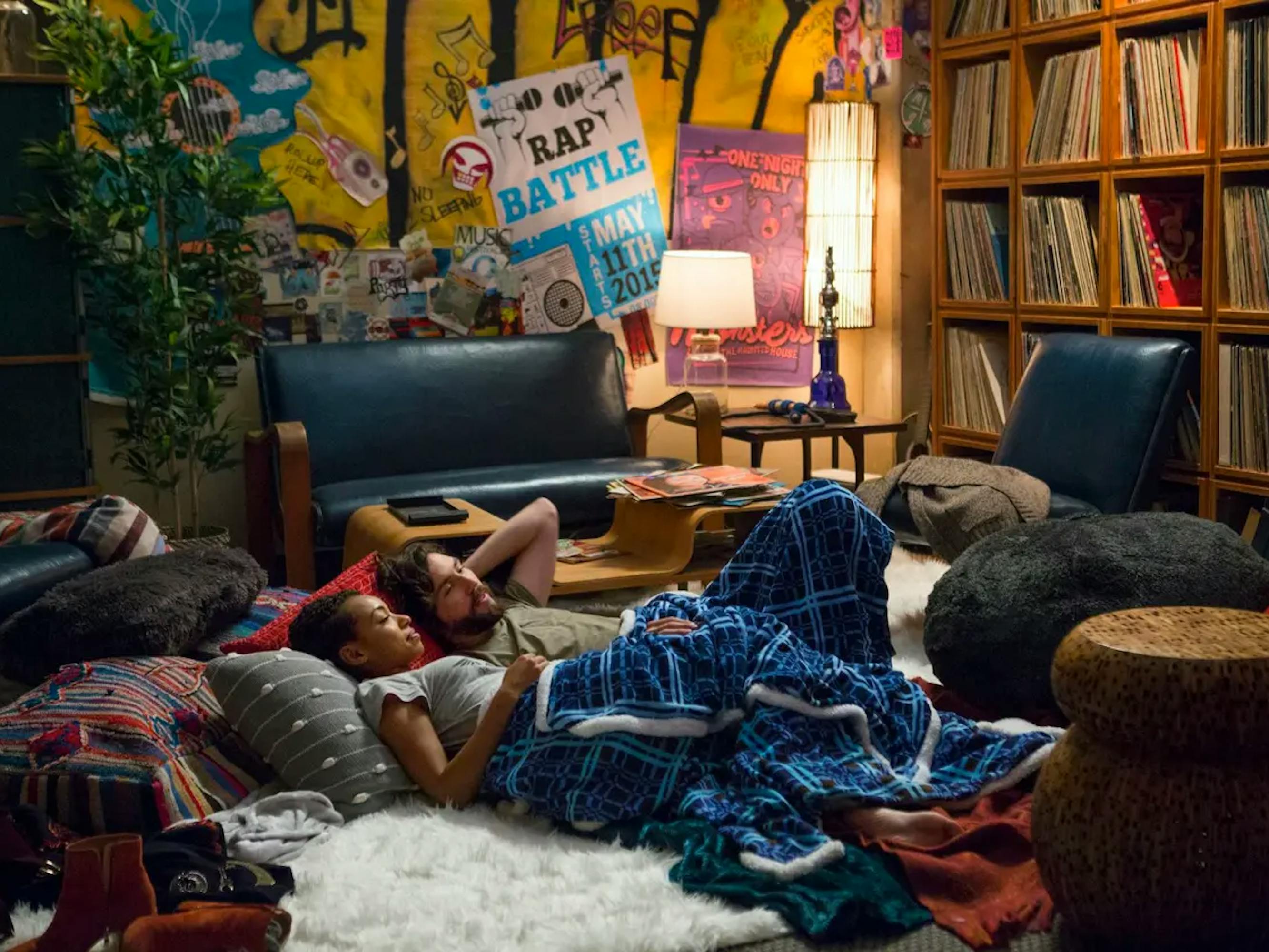 Sam White (Logan Browning) and Gabe Mitchell (John Patrick Amedori) lie on a plush white rug surrounded by chairs and pillows, and covered in a blue blanket. There are hundreds of records in shelves on the wall.