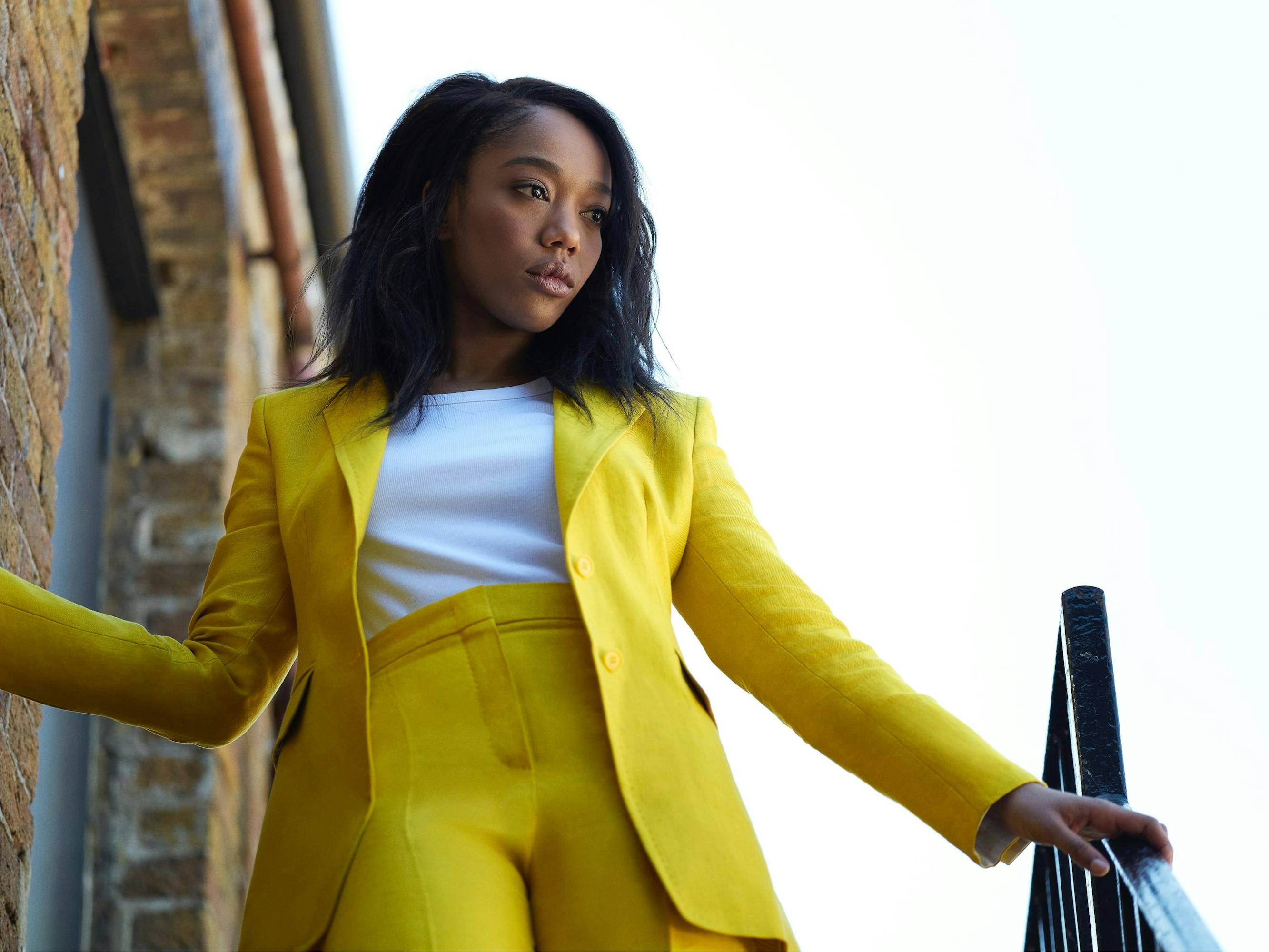 Naomi Ackie wears a vibrant yellow suit with a white top.  She stands at the top of some steps outside a brick building, her hand on the railing, gazing downwards