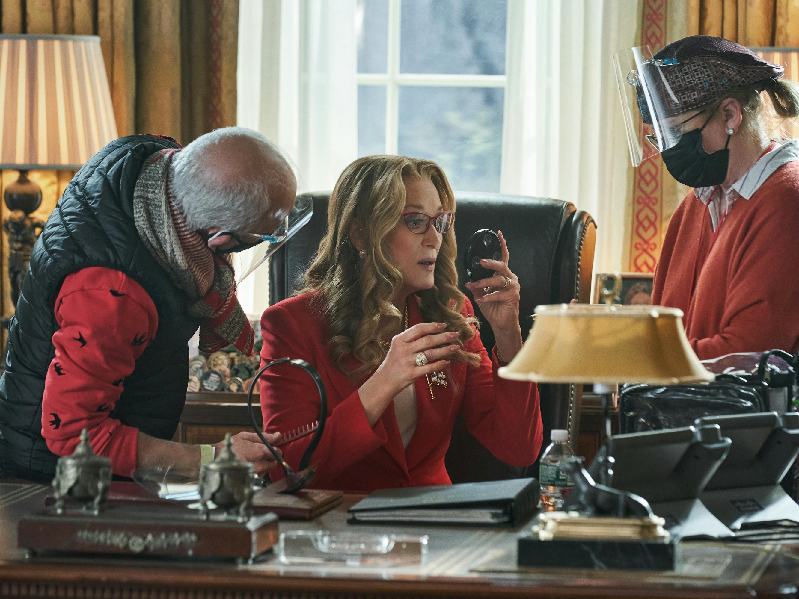 Meryl Streep and Don't Look Up crew members assemble around her oval office desk. She wears a red jacket and looks at herself in a small mirror. The crew members wear red sweaters and protective gear.