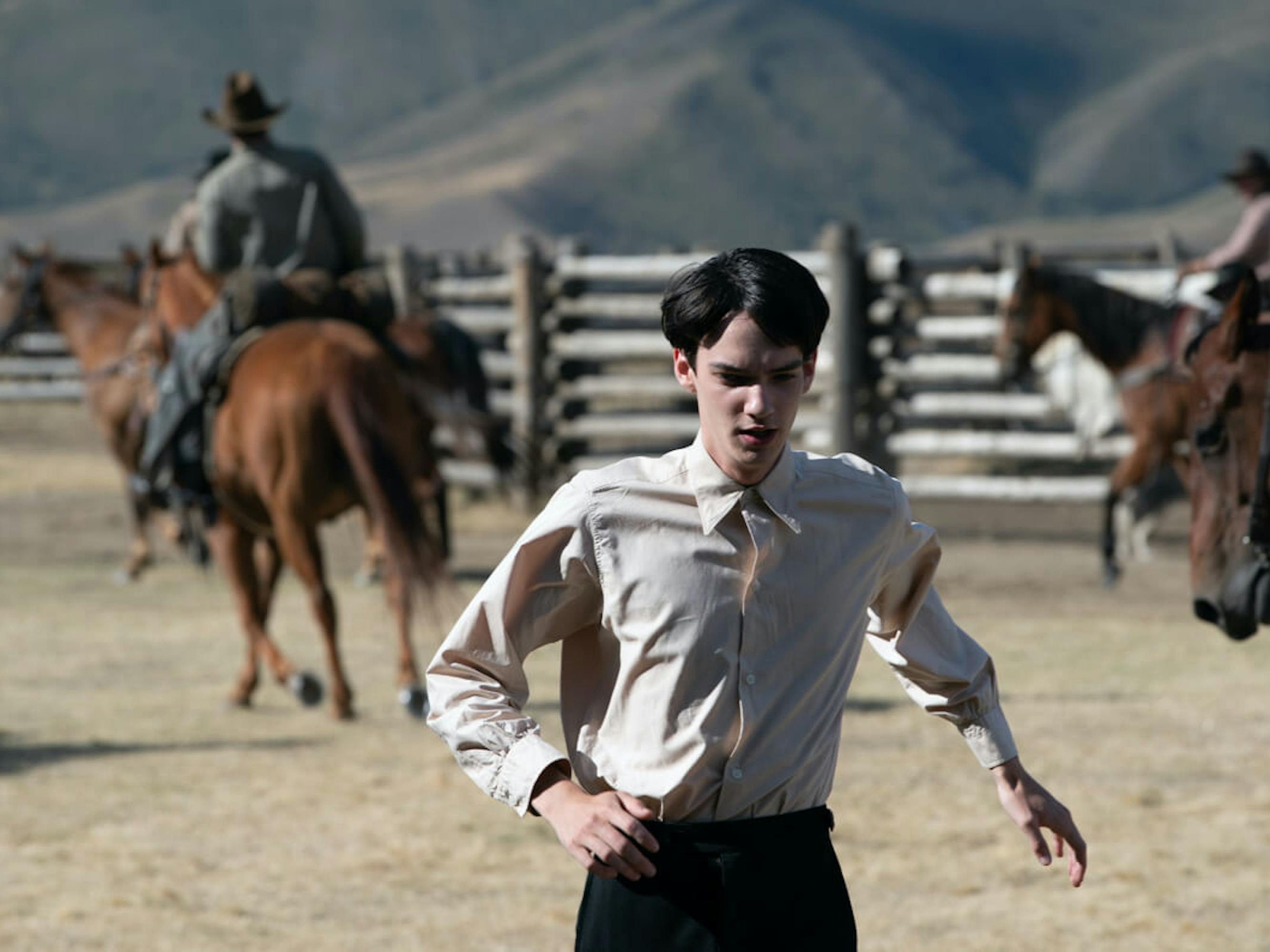 Peter (Kodi Smit-McPhee) wears a white shirt and dark pants as he runs in a fenced in area. In the background are expansive mountains, and men riding beautiful brown horses.