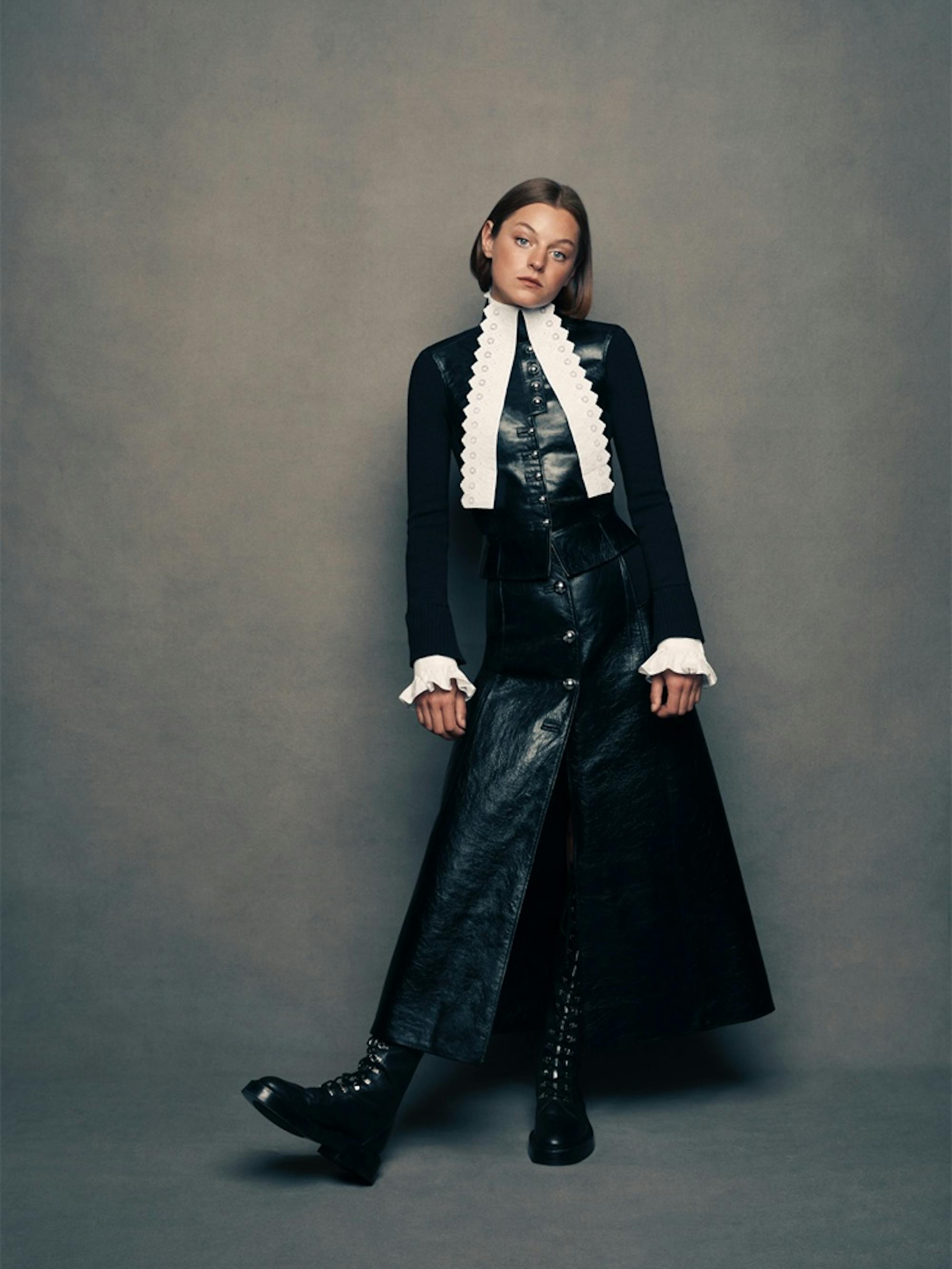 Emma Corrin against the same slate-gray background, wearing chunky black boots, a black leather vest with white-lace detail at the neck, and a matching long skirt with silver buttons. She leans casually to her left side.