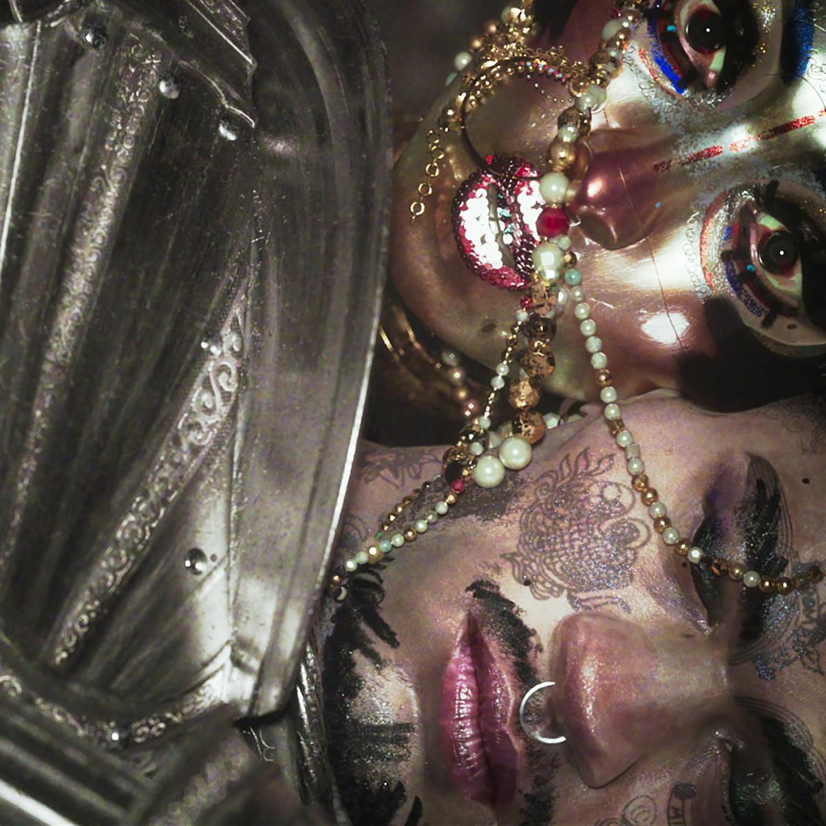 A soldier with a tattooed face and nose ring rests against a siren with a glittery painted face. Across their faces are a string of beads, and a plate of armour.