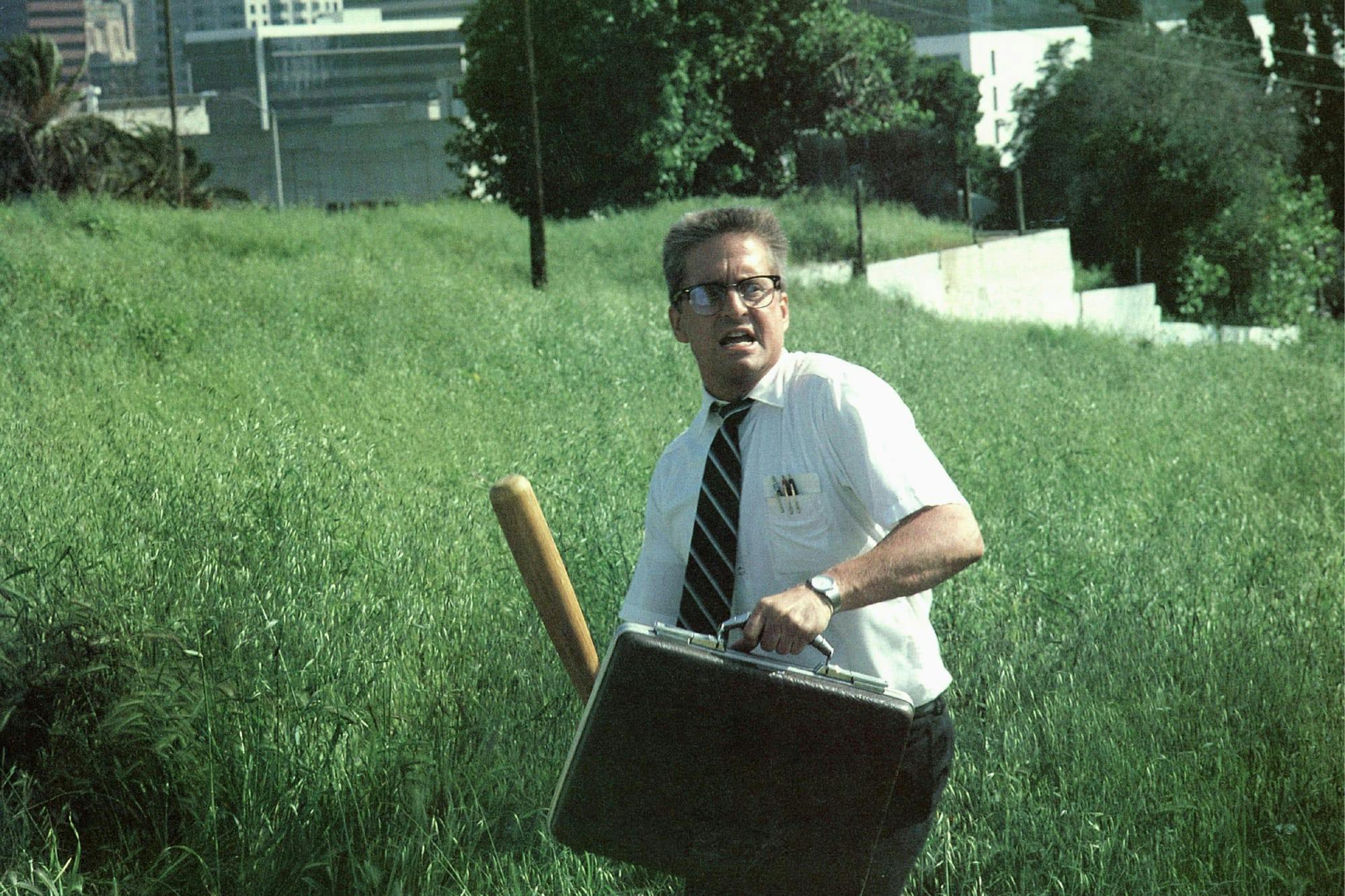 D-Fens (Michael Douglas) in Falling Down stands in a grassy field, holding a briefcase and a baseball bat.