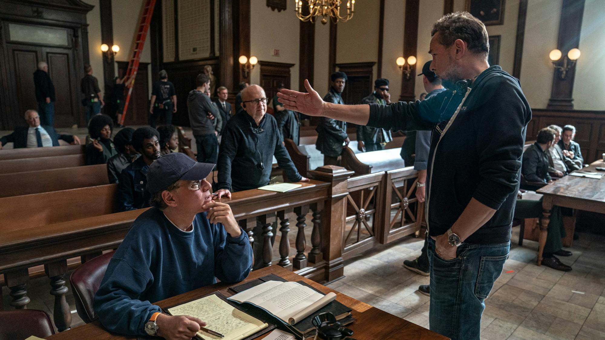 Sorkin sits at the prosecutors’ table with a notepad and the script in front of him. He looks up at Papamichael who is standing in front of the table and indicating something with his right arm. Behind them actors and crew mill about in the court gallery. The room is characterized by dark, glistening wood, gold light fixtures, and gleaming sun pouring in through tall windows.