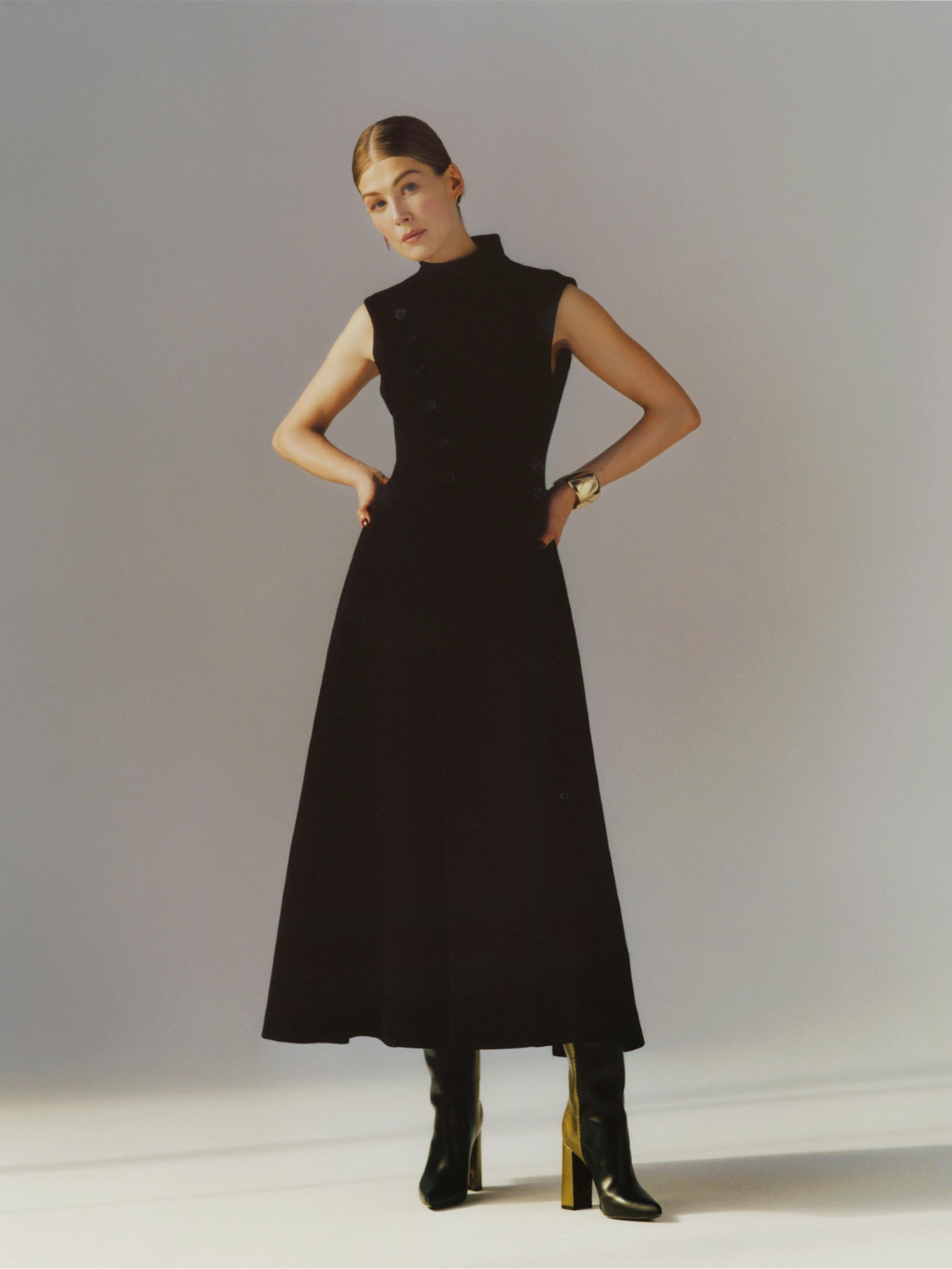 Rosamund Pike poses against a gray and white backdrop in a high-neck, long black dress and heeled black boots, her hands on her hips.