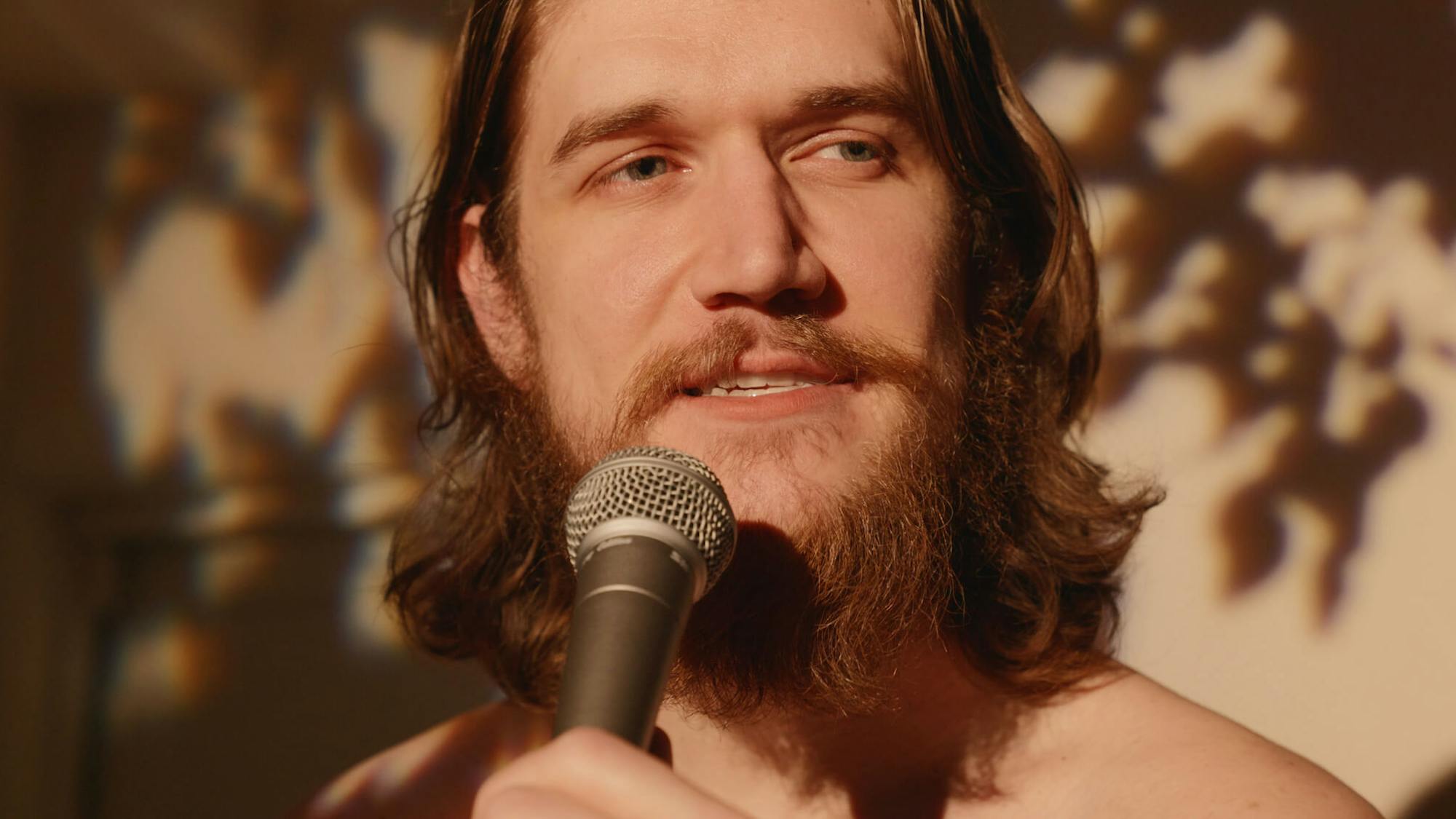 In this close up shot, Bo Burnham sings into a microphone. He is lit by dappled light, his hair is long, and he has a beard and mustache.