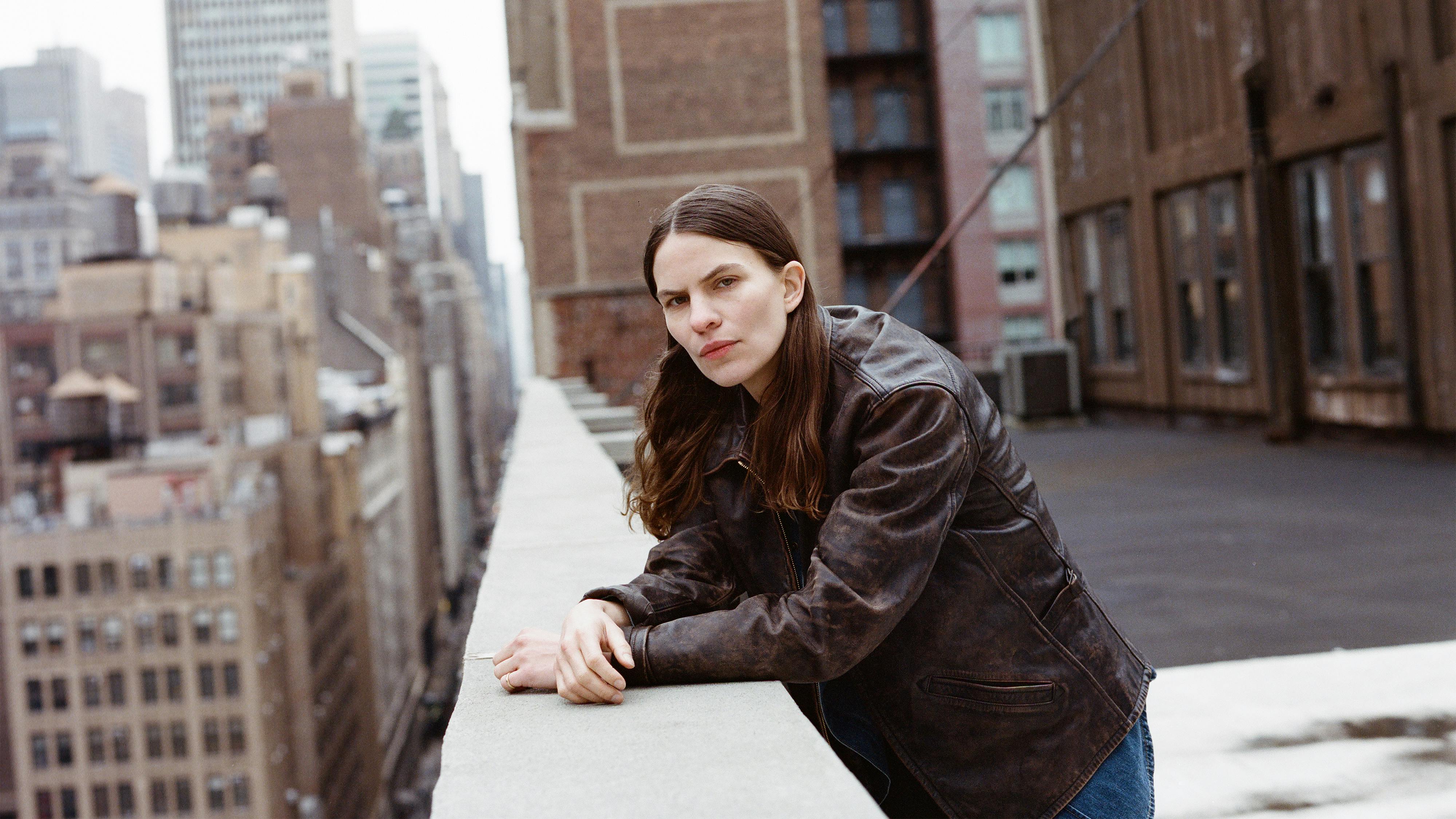 Eliot Sumner wears a brown leather jacket and leans on a roof wall overlooking New York City.