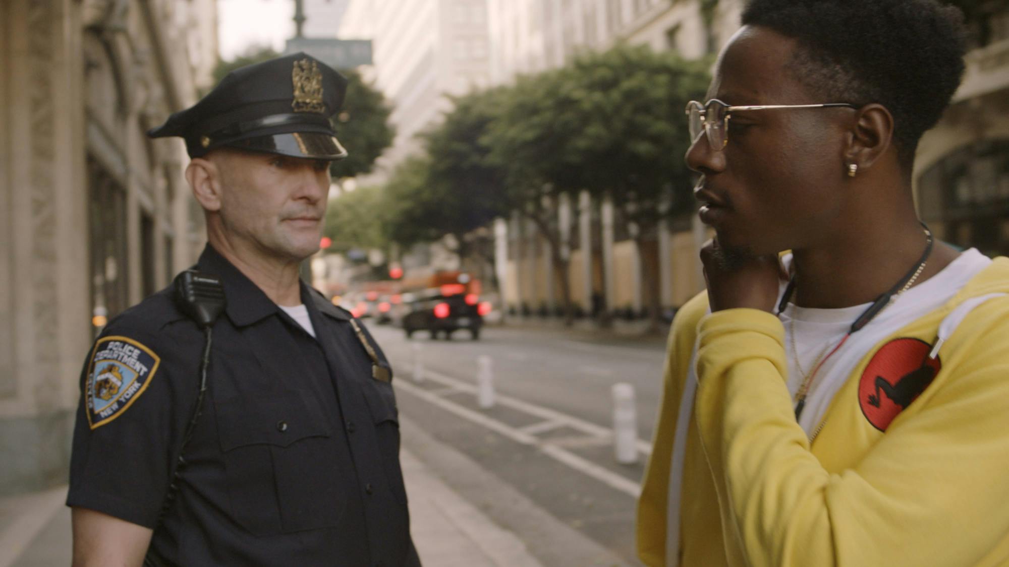 Howard and Bada$$ look at each other on the street. Howard is in uniform, and his posture is threatening. Bada$$ wears his yellow hoodie and white shirt, and is edging away. In the background are trees and cars at a light. 