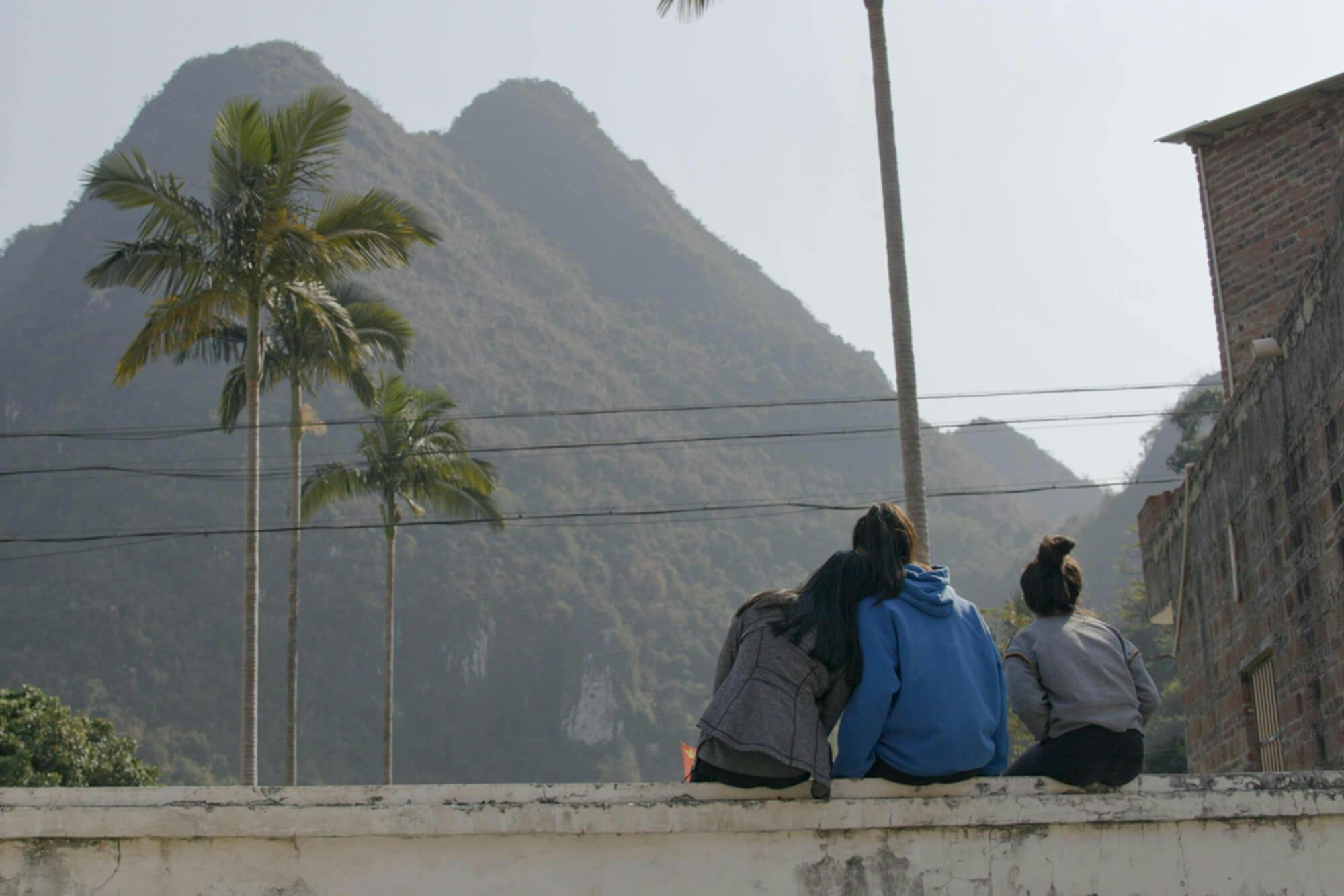 Three girls sit on a wall looking off at two mountain peaks. There are a few palm trees in the foreground. 