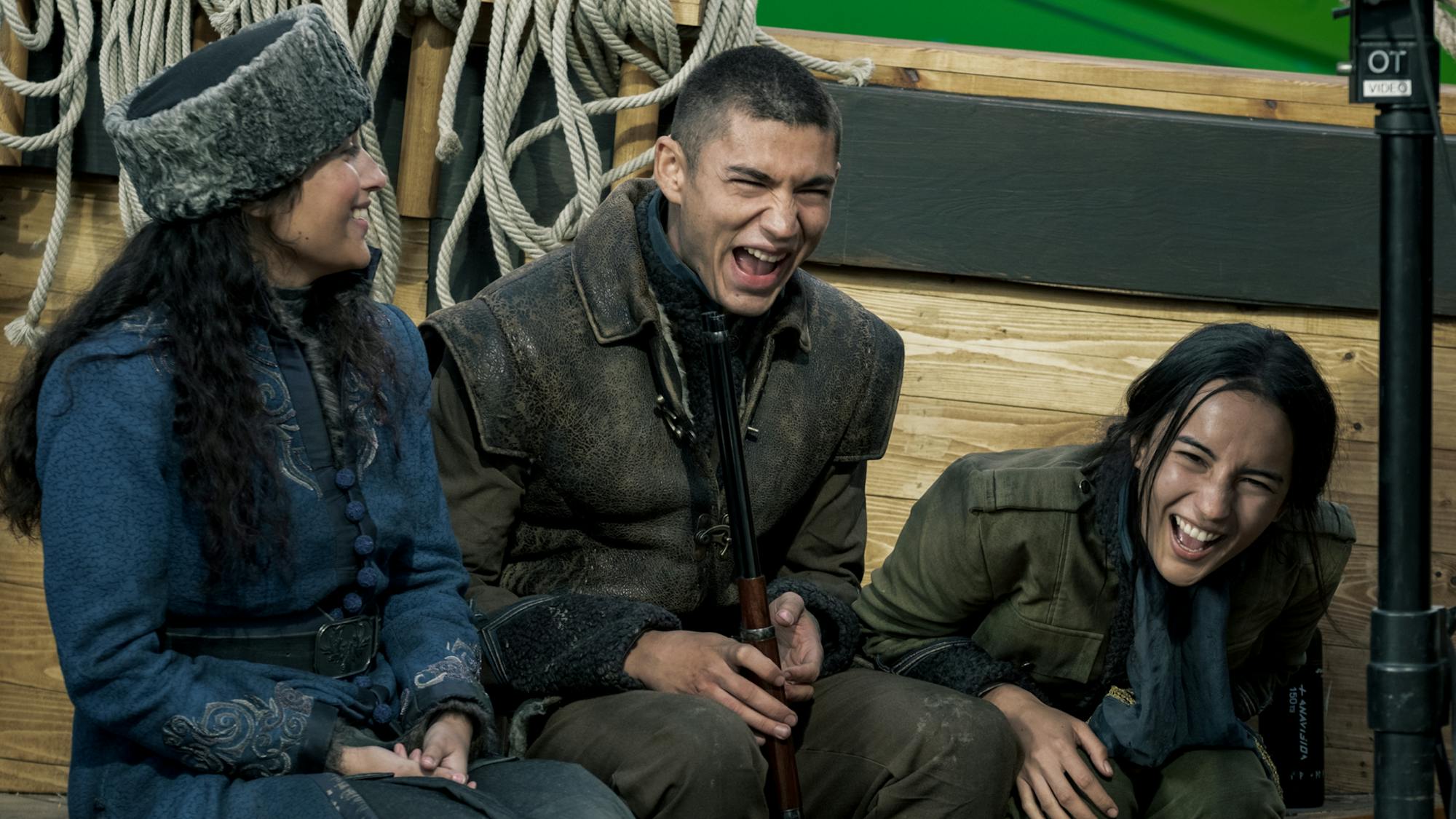 Sujaya Dasgupta, Archie Renaux, and Alina Starkov sit together. Dasgupta wears a blue jacket and fur hat. Renaux wears a brown jacket and carries a gun. Mei Li wears a blue scarf and green jacket. All three are laughing uproariously.