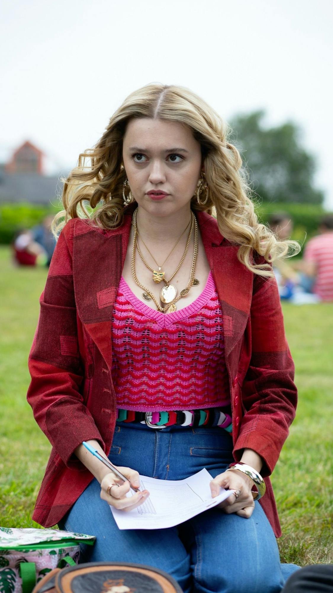 Aimee Gibbs (Aimee Lou Wood) wears a pink sweater, red coat, blue jeans, and a colorful belt. She kneels in the grass writing something on a piece of paper.