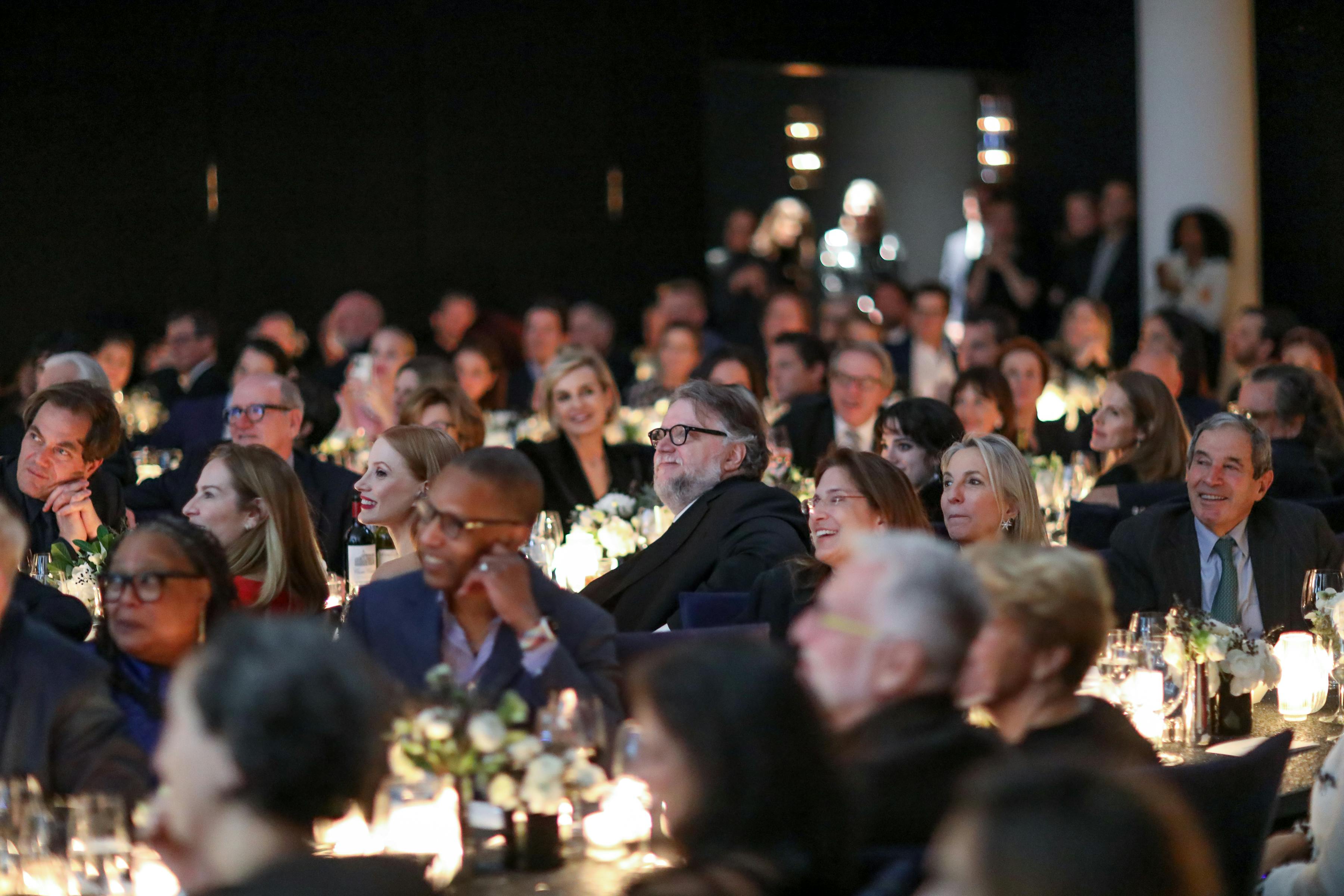 A smiling crowd of people, with Guillermo del Toro sitting at the center. The tables are alight with candles and flowers, and people are all looking at something offscreen.