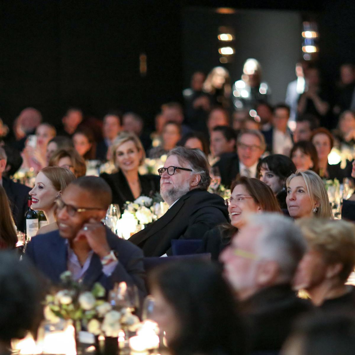 A smiling crowd of people, with Guillermo del Toro sitting at the center. The tables are alight with candles and flowers, and people are all looking at something offscreen.