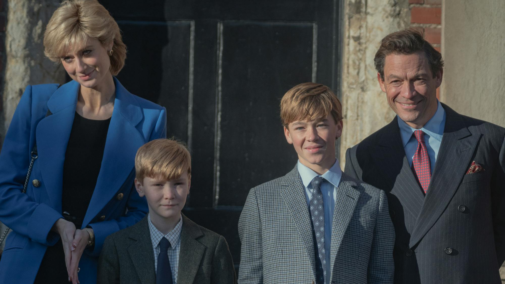 Princess Diana (Elizabeth Debicki), Prince Harry (Will Powell), Prince William (Senan West), and Prince Charles (Dominic West) stand together smiling slightly. Diana wears a blue jacket, and the boys wear navy ties and checkered jackets. Charles wears a double breasted jacket and red tie.