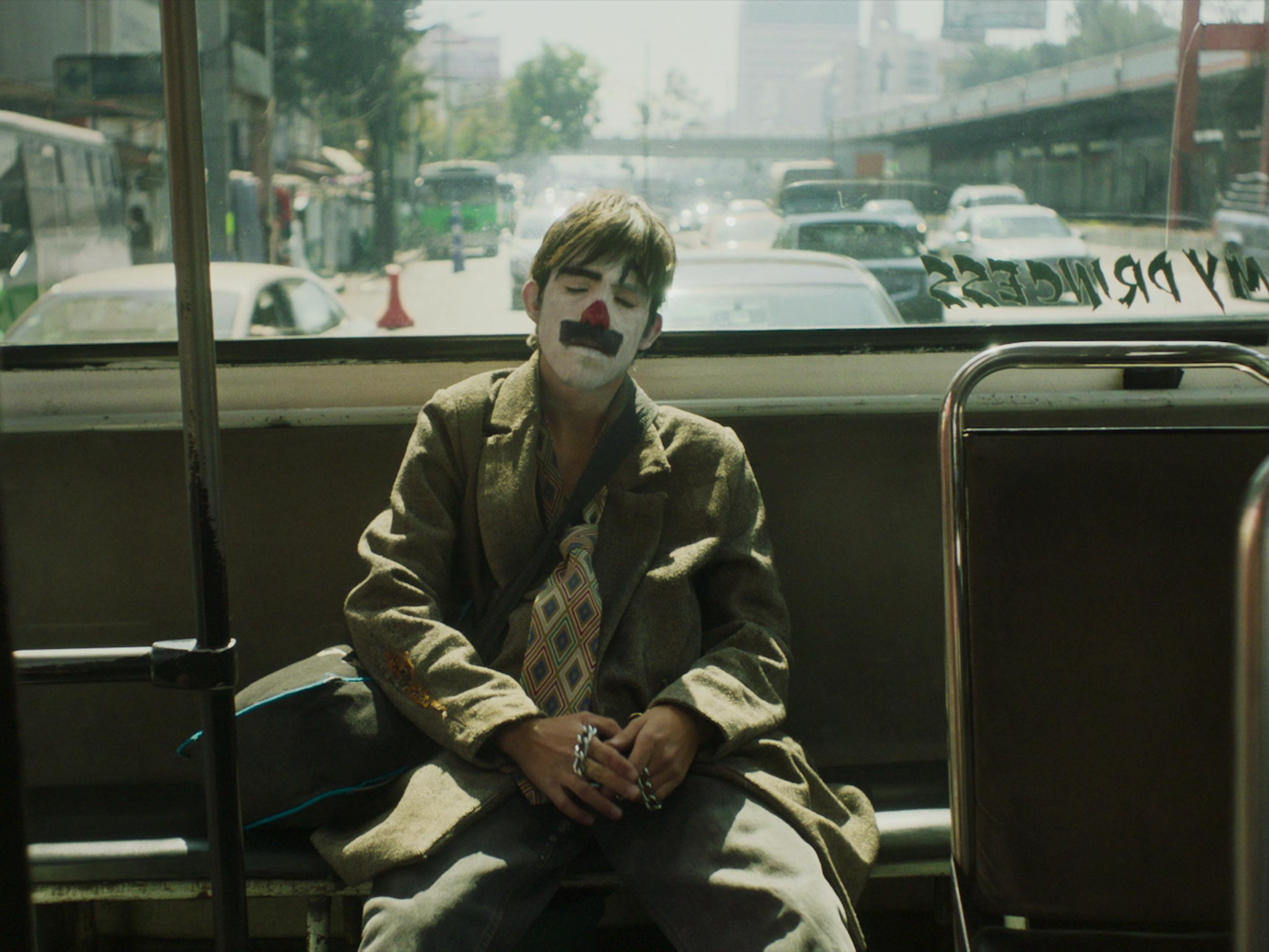A character from Chicuarotes sits in the back of the bus in a joker-esque outfit.