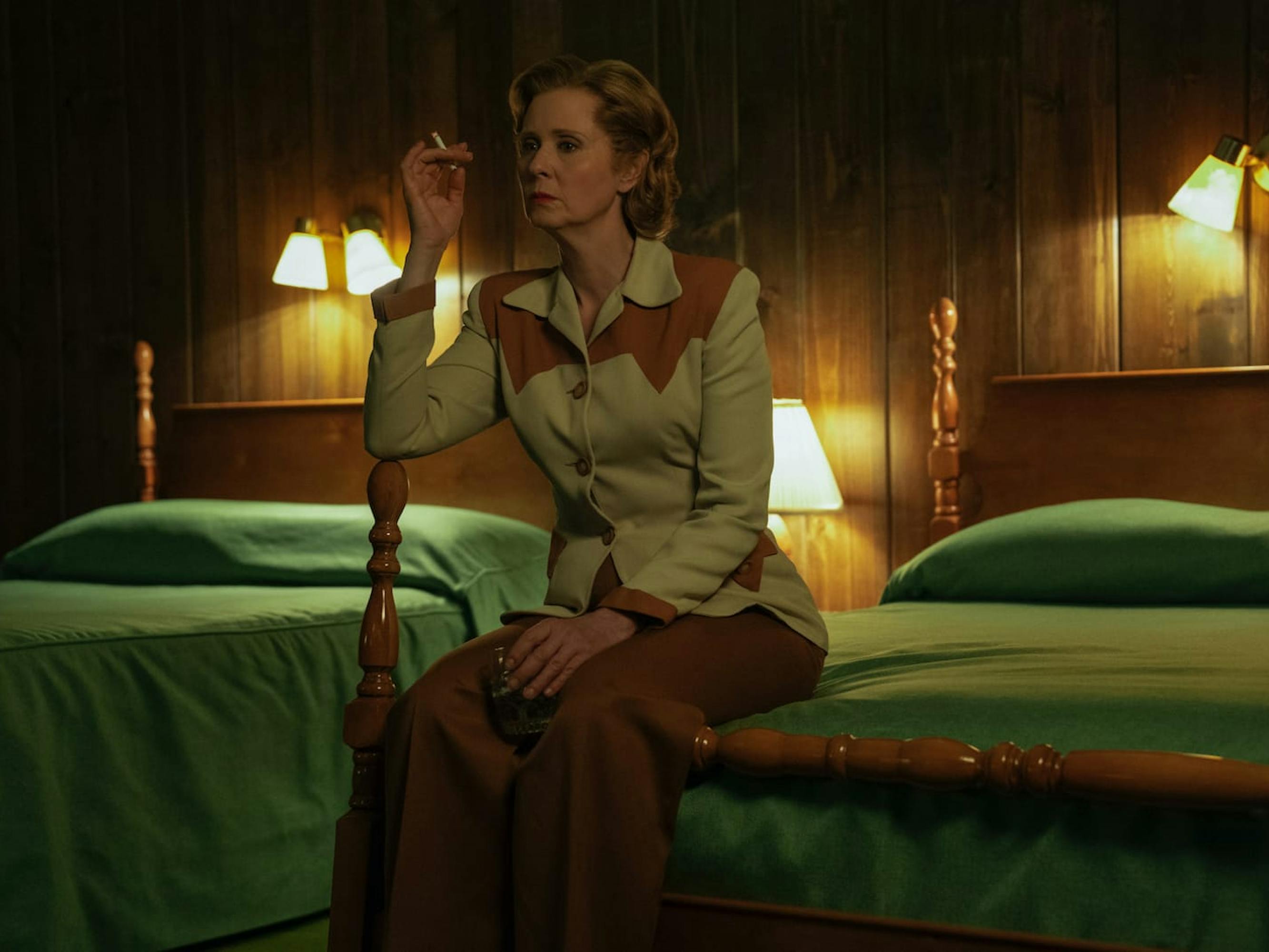 Gwendolyn Briggs sits on a four poster bed smoking a cigarette and drinking out of a glass. The sheets are green, and the room is lit by 5 bright lamps. She looks stressed although her chic western outfit says otherwise.