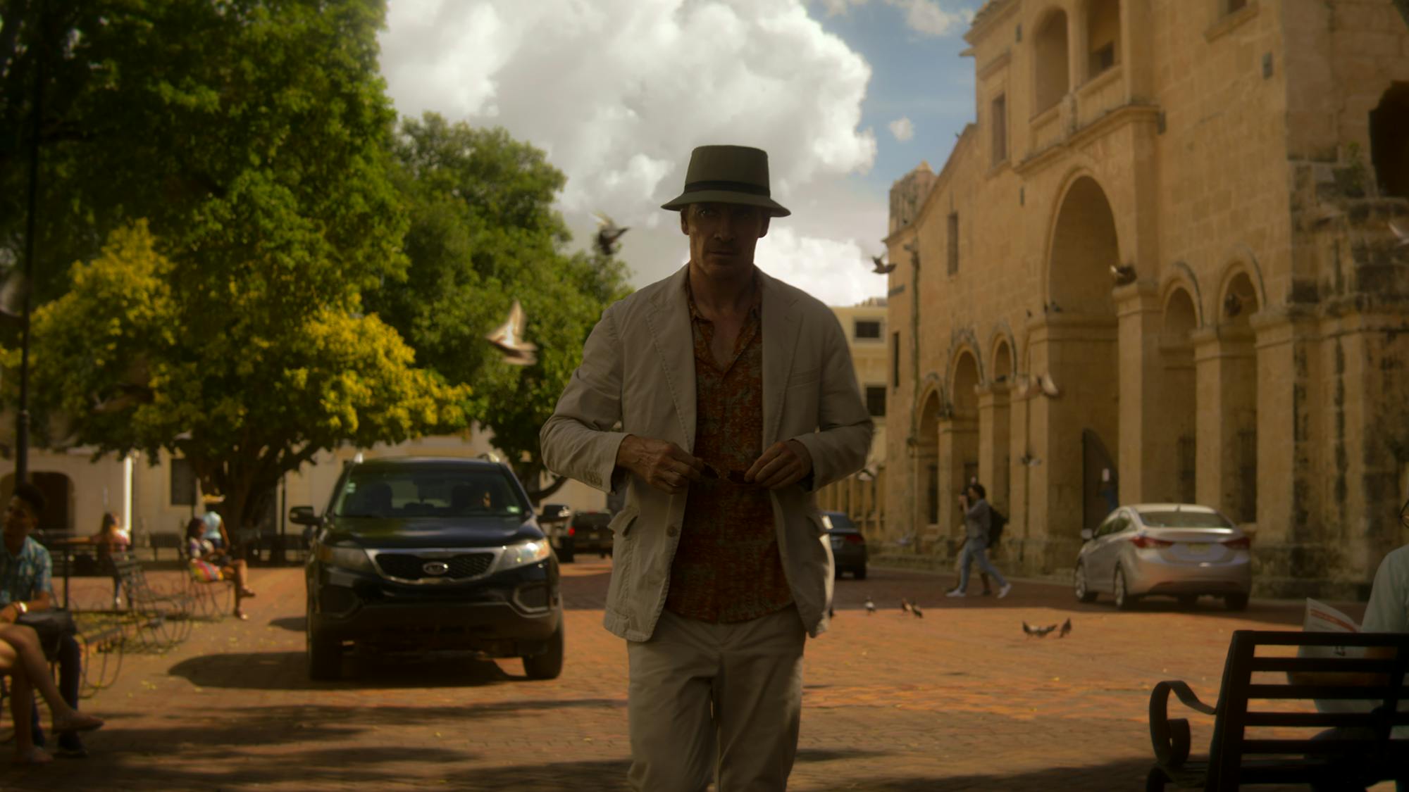 The Killer (Michael Fassbender) walks through a sunny scene in a tan suit and a bucket hat.