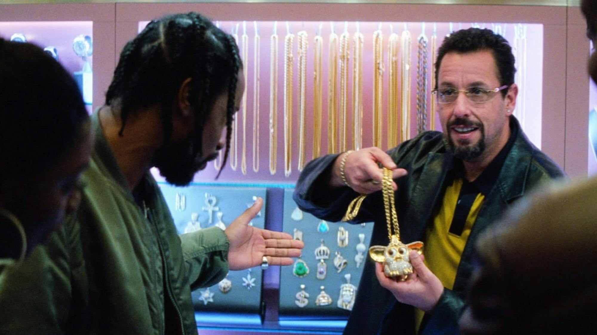 Three figures hover around Adam Sandler as he holds a gold and diamond Furbie necklace.