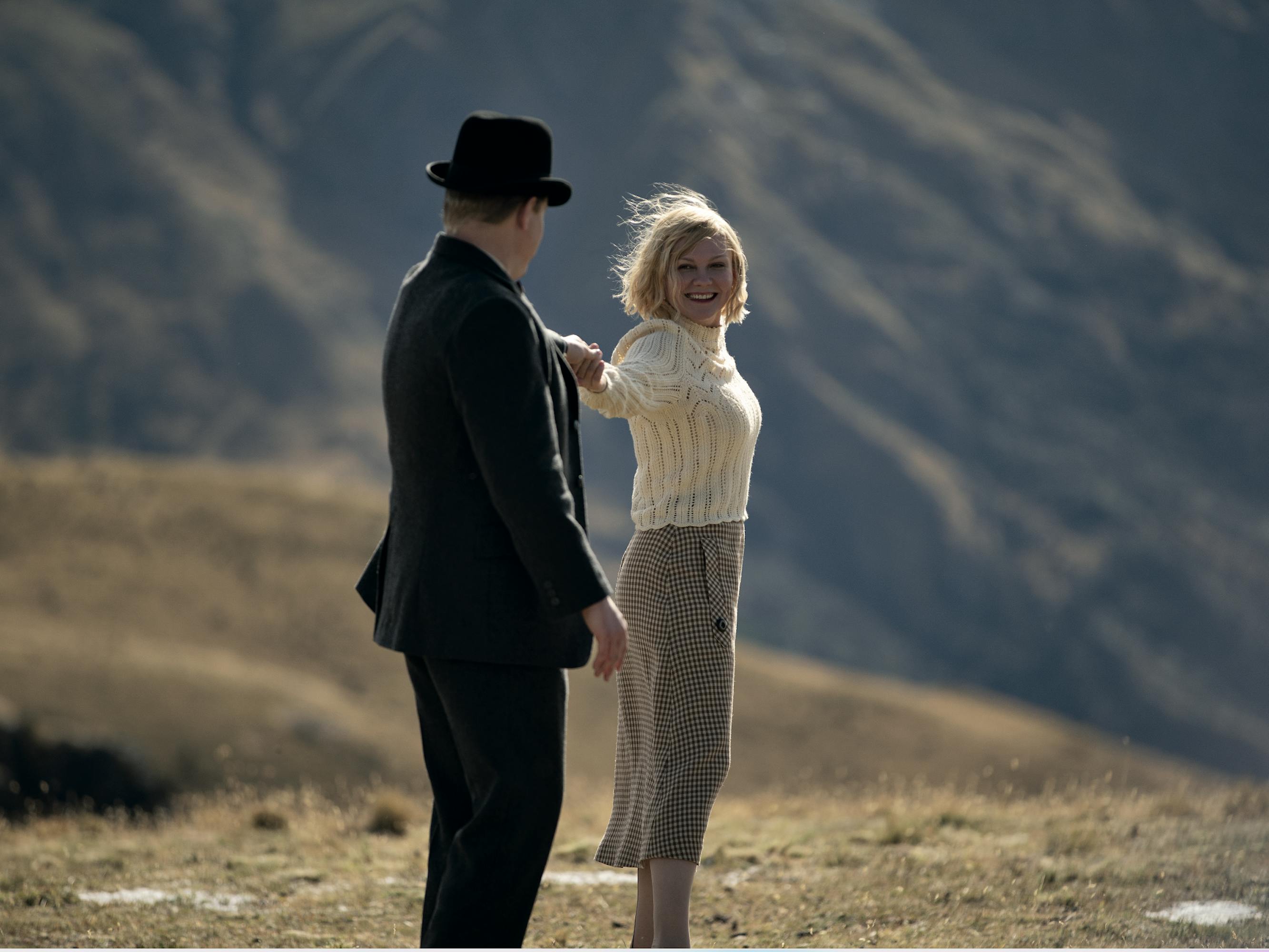 Jesse Plemons and Kirsten Dunst dance on a hilltop. He wears a dark suit and a hat, she wears a sheer blouse and a khaki skirt. The mountain behind them is blue, and the ground is dusty brown.