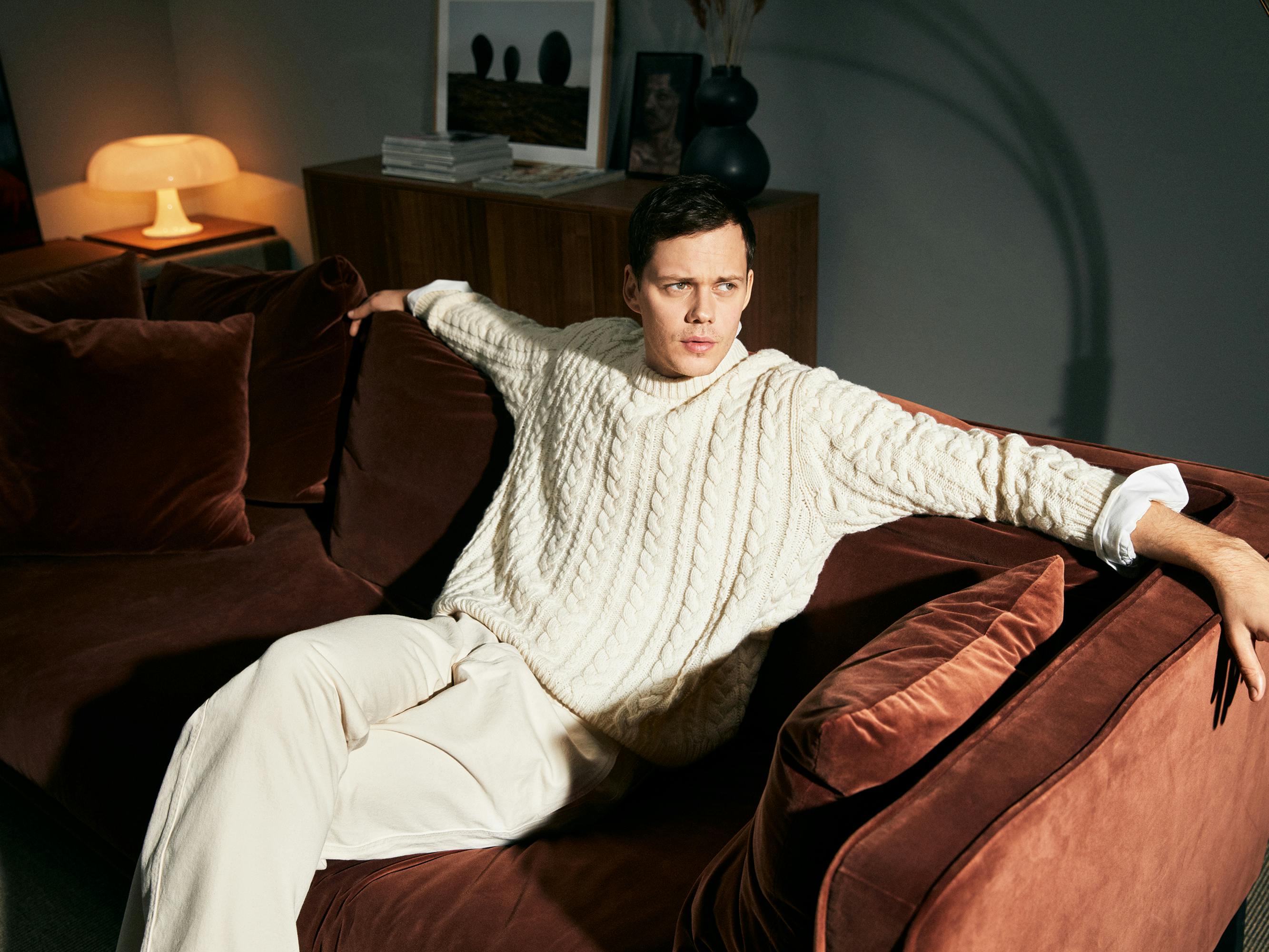 Bill Skarsgård wears a cream colored outfit and stretches out on a brown velvet couch. 