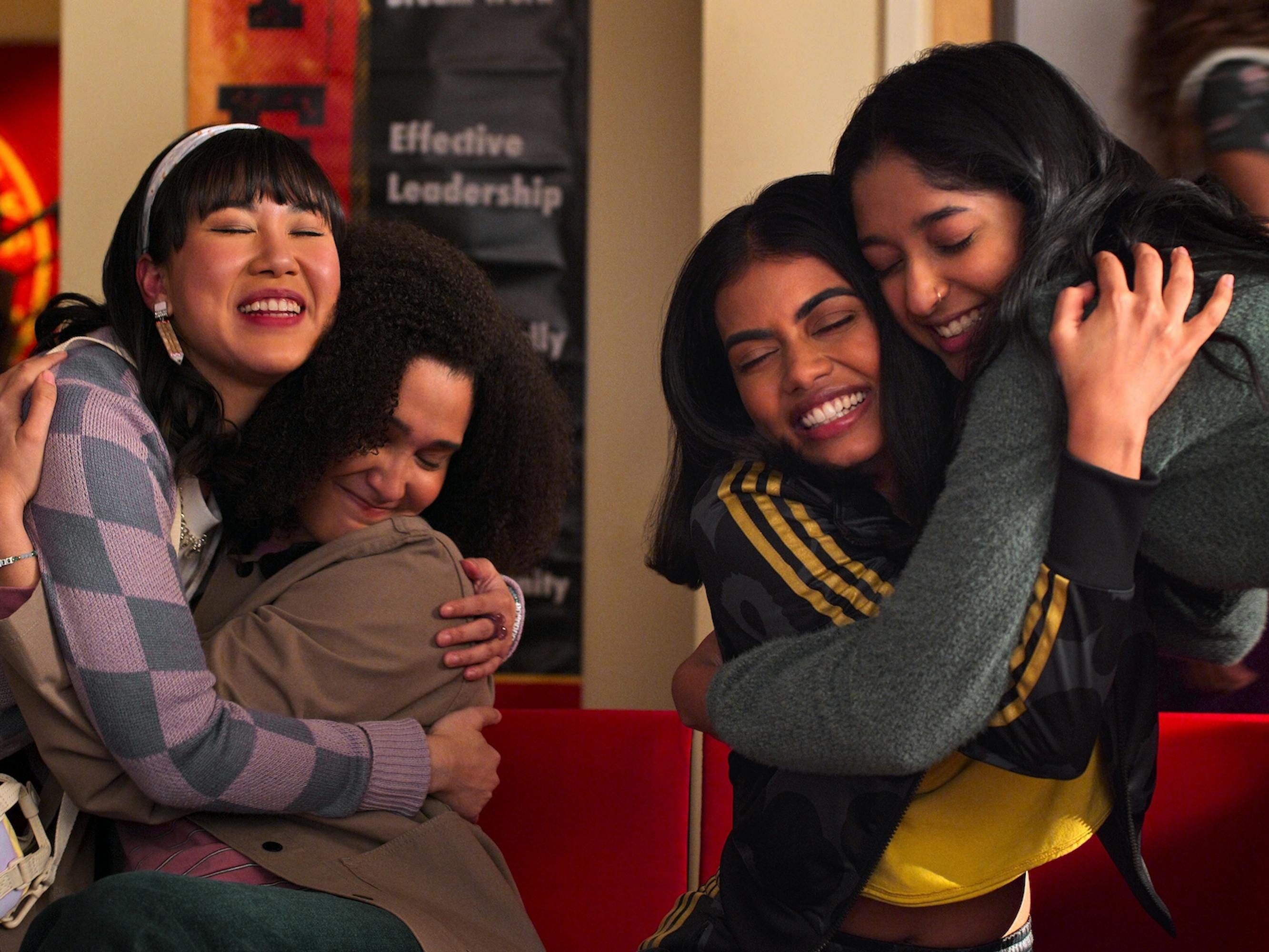 Eleanor Wong (Ramona Young), Fabiola Torres (Lee Rodriguez), Aneesa (Megan Suri), and Devi Vishwakumar (Maitreyi Ramakrishnan) hug. They're sitting on Sherman Oaks red- chairs.