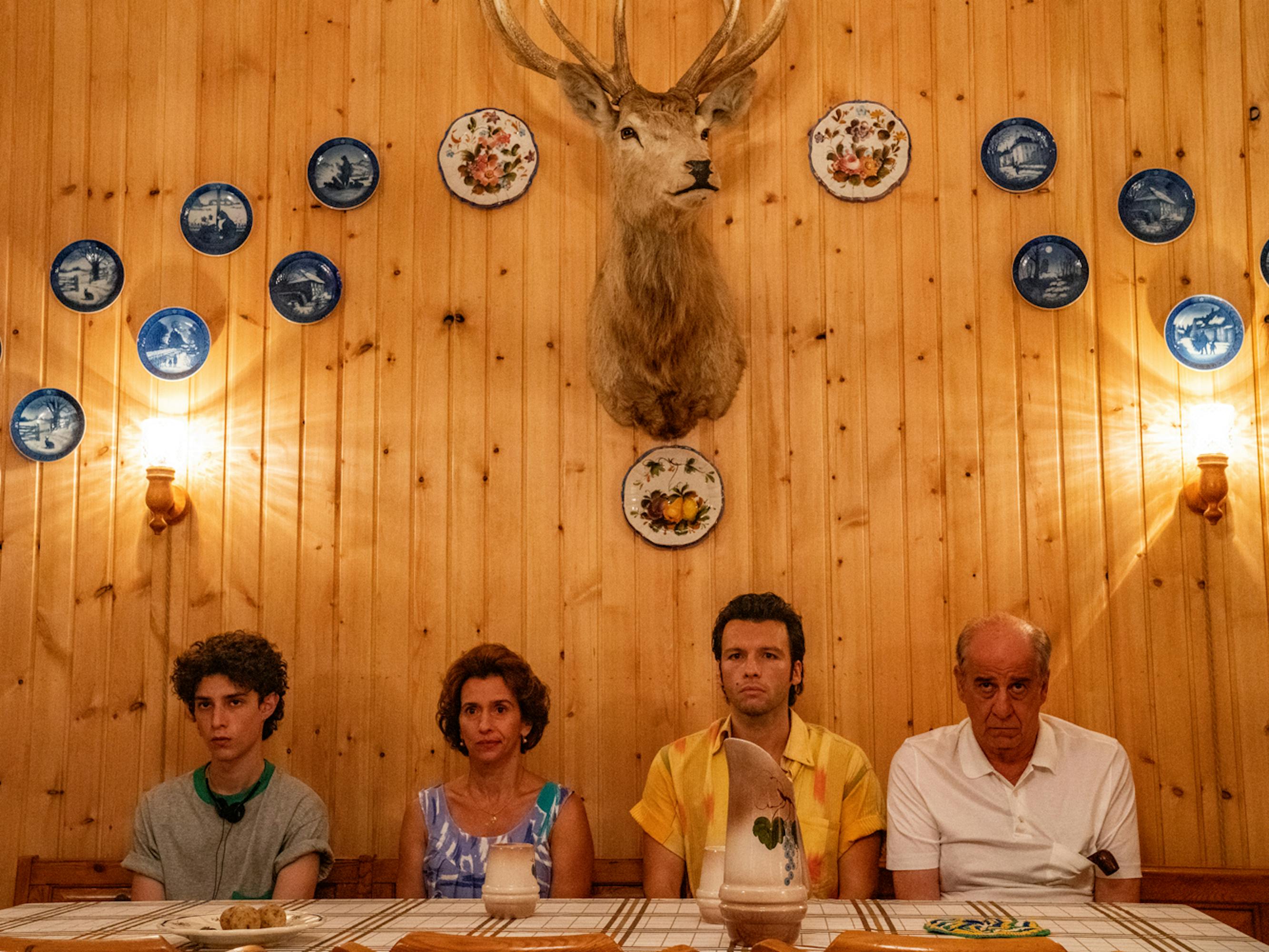 The cast of The Hand of God sits at a dinner table. Behind them is a dead animals bust and many blue plates.