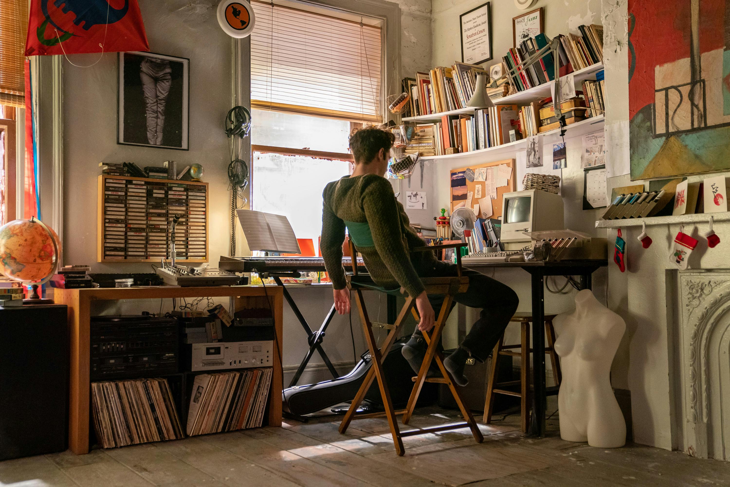 Andrew Garfield works at his desk in a crowded sunlit room. The walls are lined with books, CDs, and colorful posters.Garfield sits in a green and wood chair and leans back. Near his right knee is a bust of a woman's top.