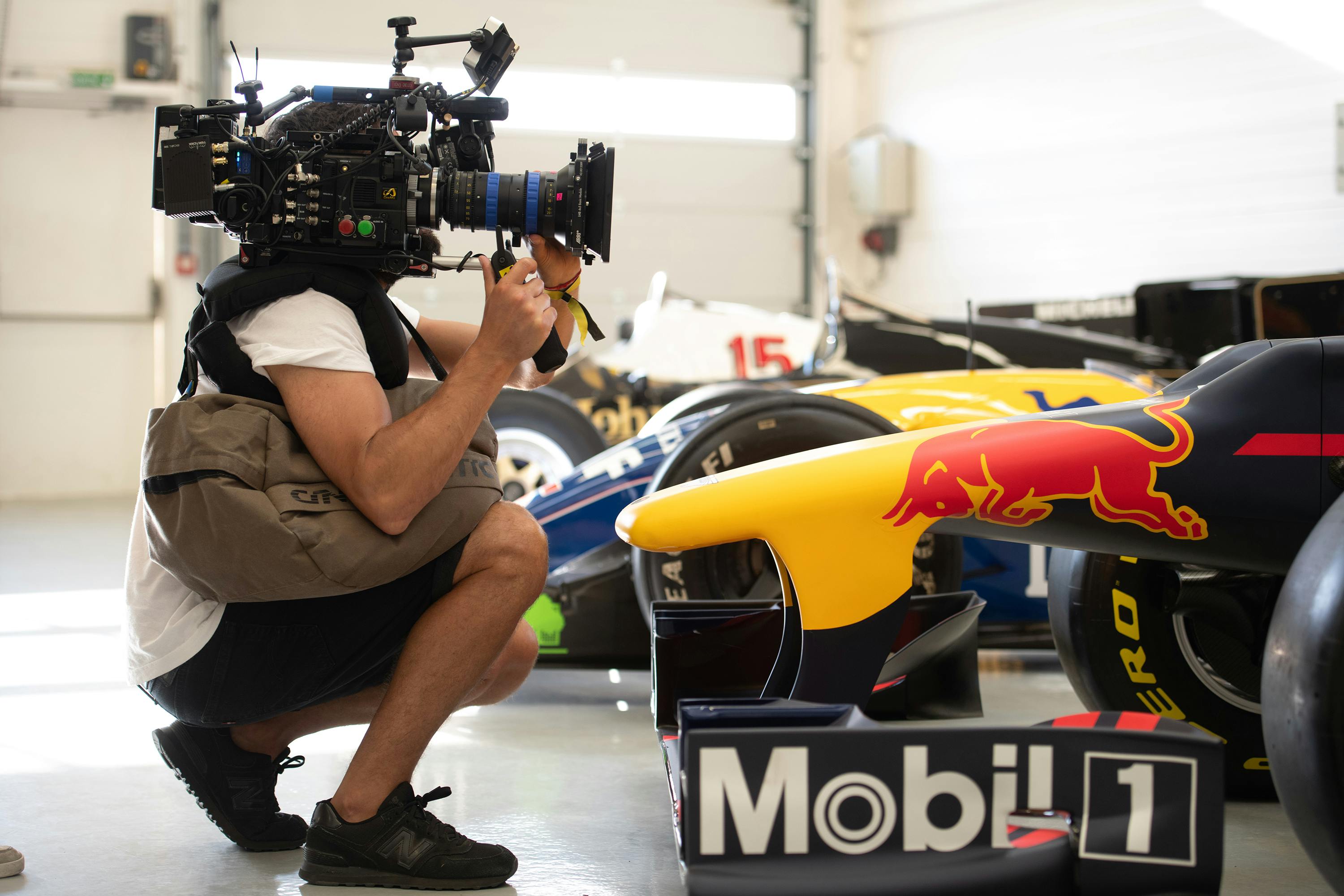 Behind the scenes of Formula 1: Drive to Survive. A person holding a camera crouches next to a yellow, red, and black car with Mobil 1 on its side.