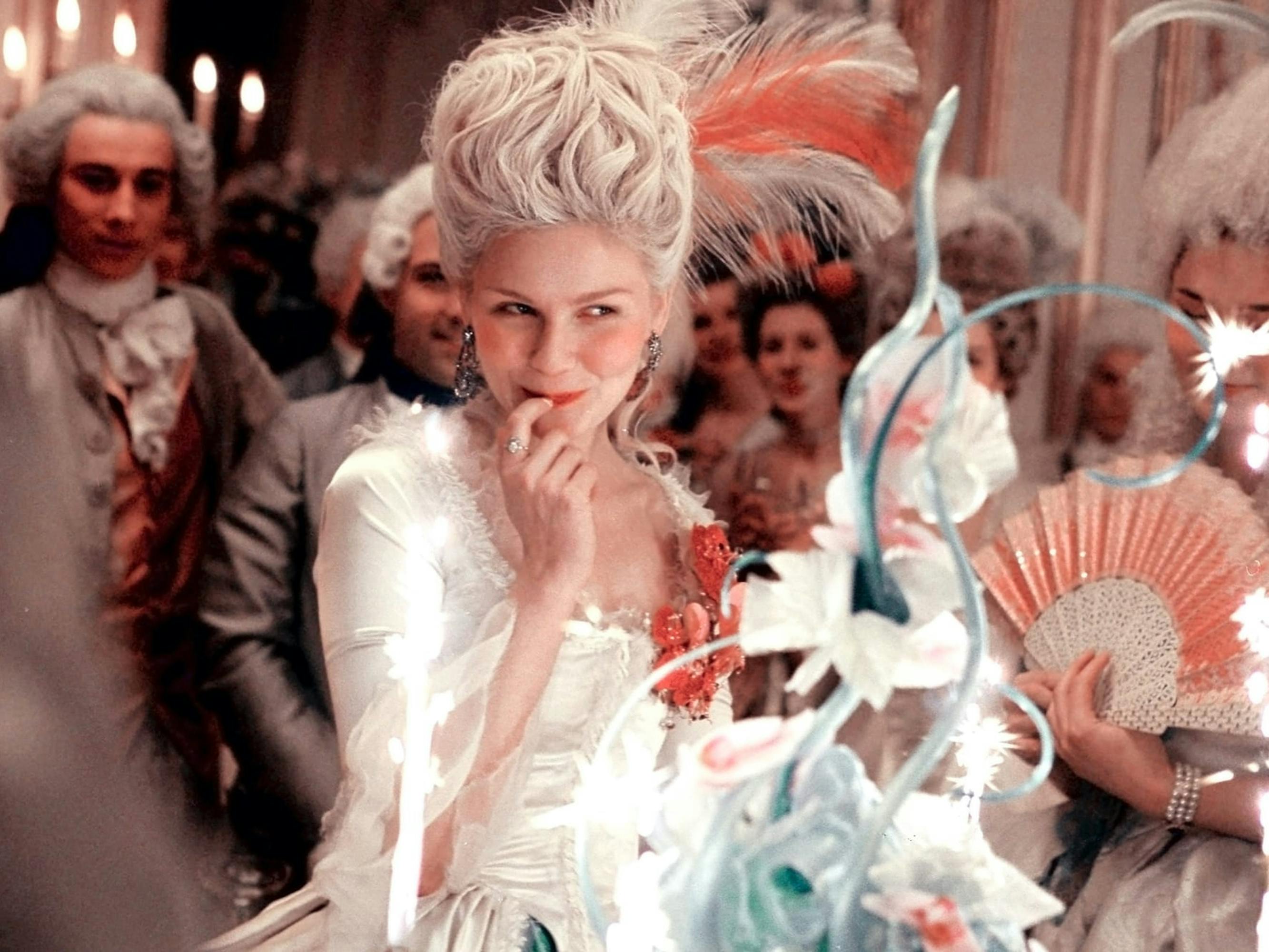 Marie Antoinette (Kirsten Dunst) wears her hair high and a silky white dress. She’s surrounded by many others in similar garb.