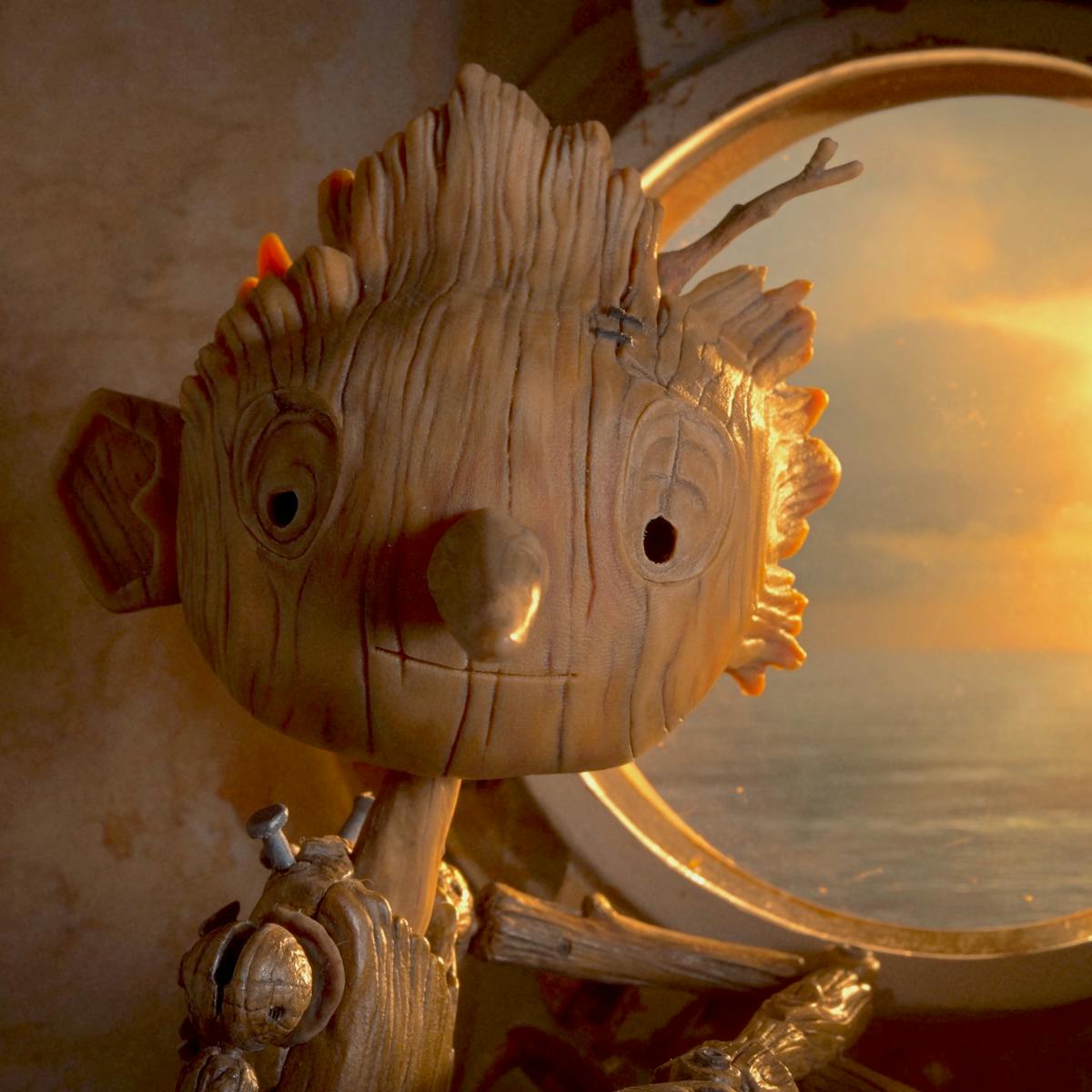 Pinocchio (Gregory Mann) looks out of a circular window at the ocean and an orange setting sun. 