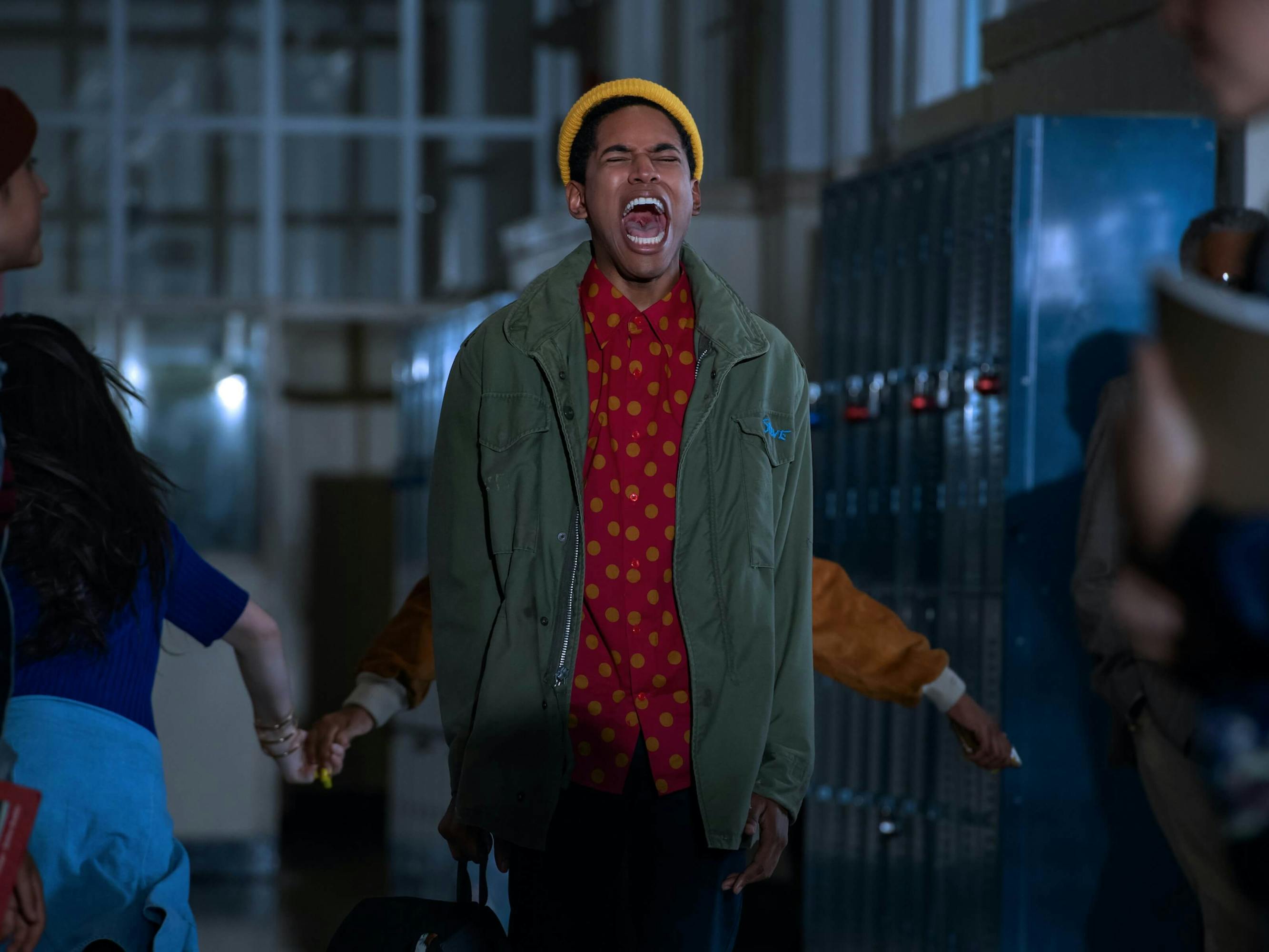 Harrison as Steven Harmon in Monster. He wears a red buttoned-down shirt, a green jacket, jeans, and a yellow beanie. Other kids stand in the hallways facing different directions. You can tell it’s a school by the lockers to his right.