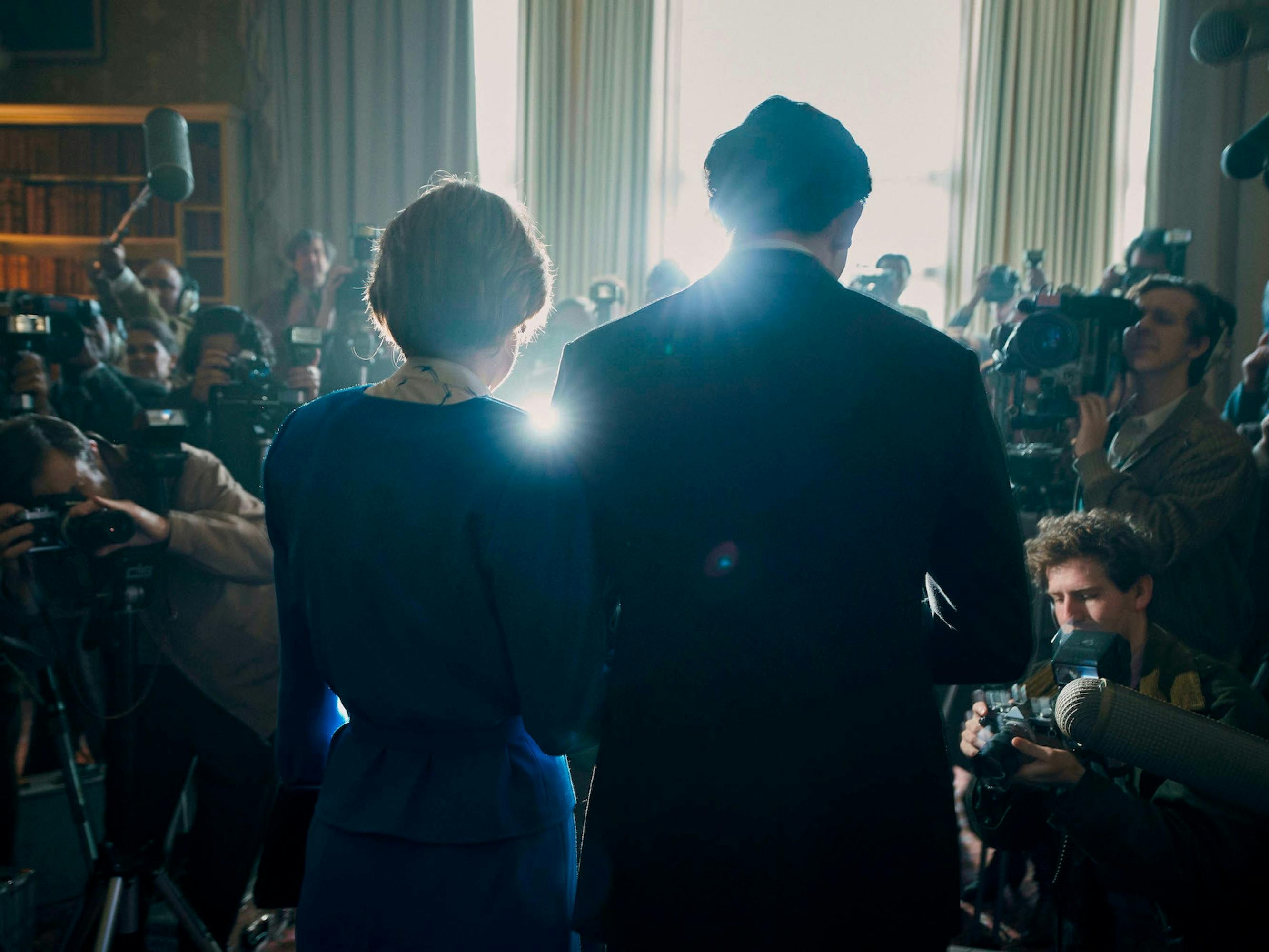 Lady Diana Spencer (Emma Corrin) and Prince Charles (Josh O’Connor) stand with their backs to us. Their figures are lit by paparazzi’s flashes.